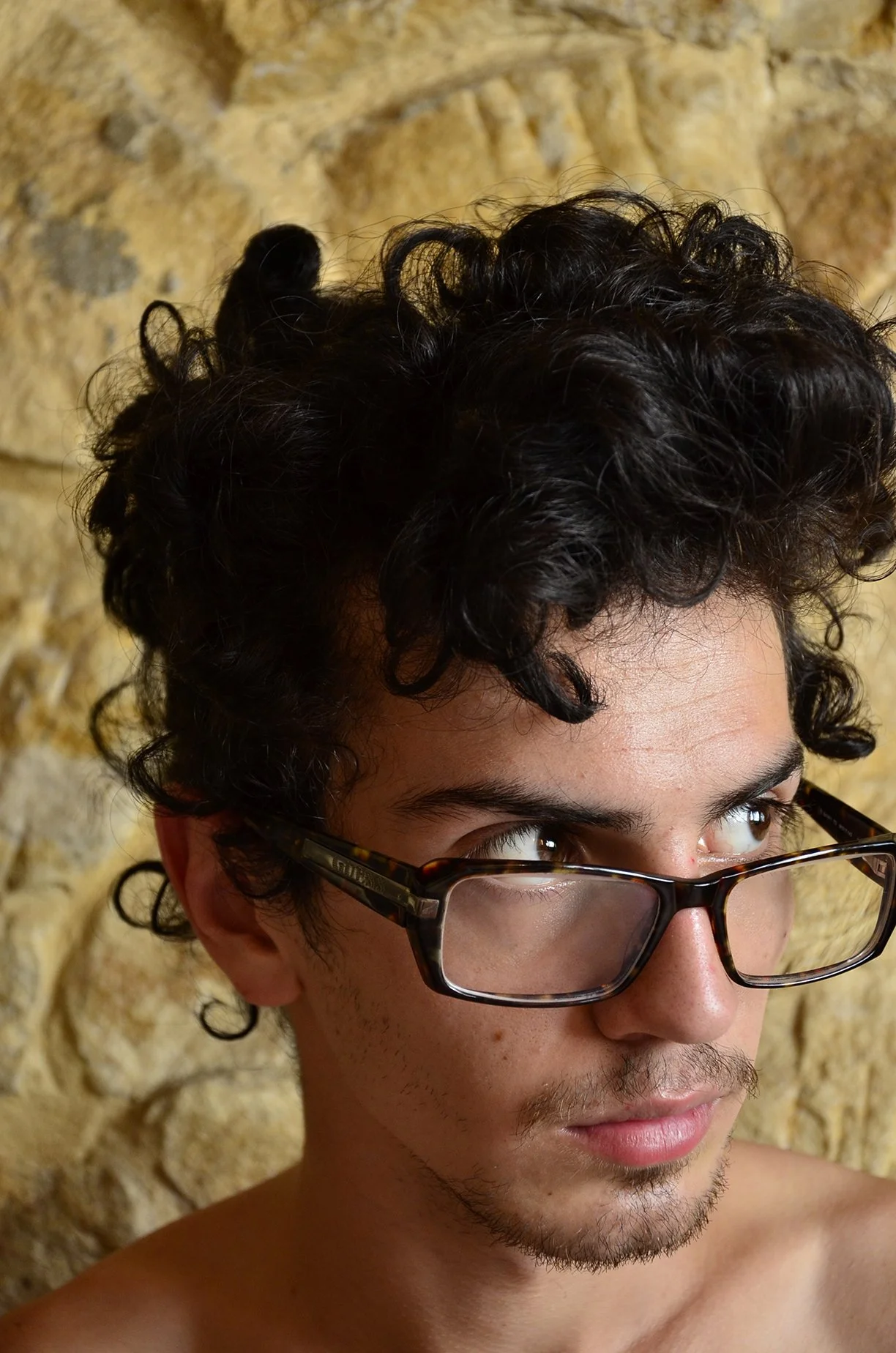 A young man with curly dark hair and glasses looking to the right in front of a stone wall.