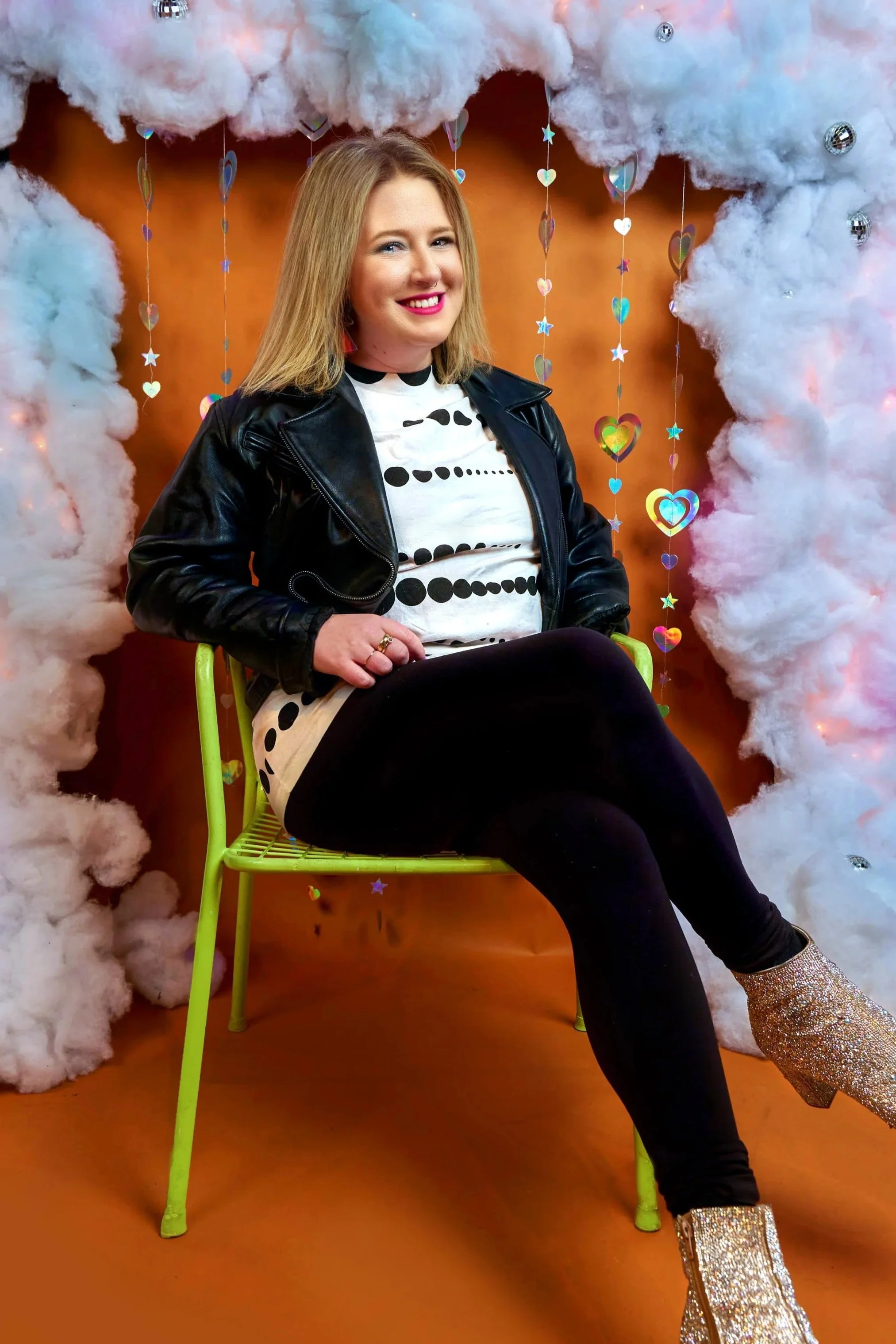 A woman sitting on a lime green chair in front of a decorative backdrop with fluffy white clouds, hanging holographic hearts, stars, and small mirror ornaments, smiling at the camera.