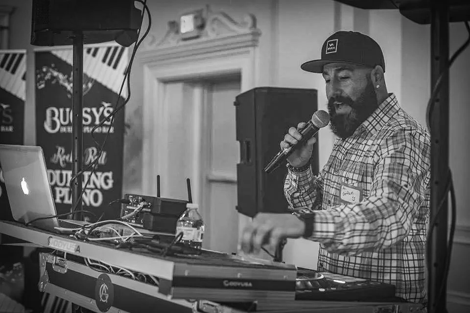 A man DJing using a laptop and turntables at an indoor event, speaking into a microphone, wearing a plaid shirt and a baseball cap.