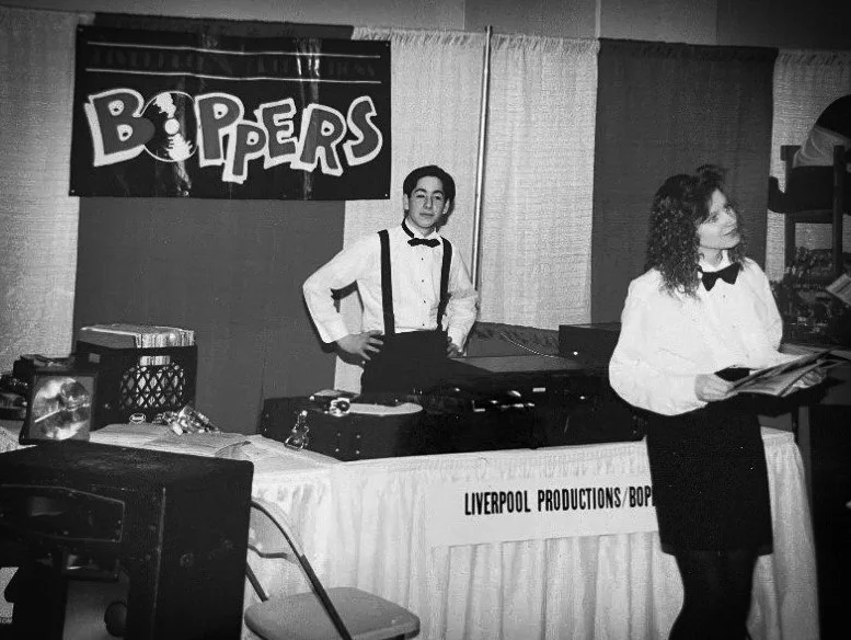 A black and white photo of two women in formal attire with DJ equipment and a Liverpool Productions banner at a 1970s event.