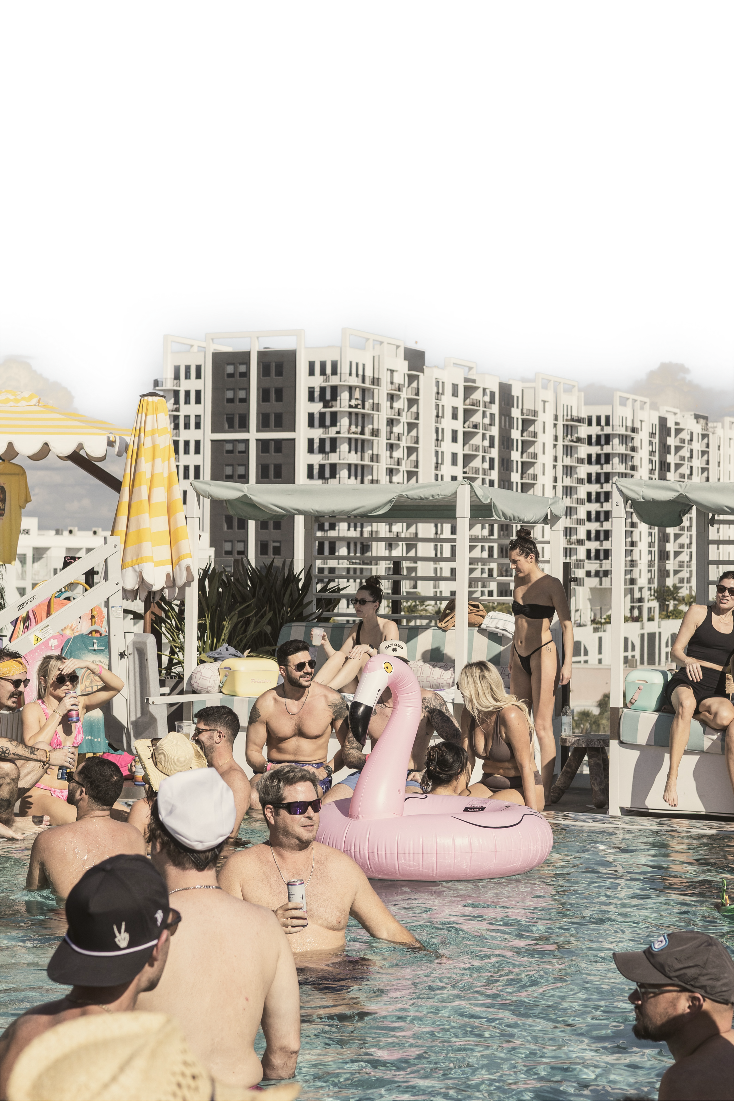 People relaxing and socializing by a rooftop pool with high-rise apartment buildings in the background, some wearing swimwear, sunglasses, and hats, with pool floats and umbrellas.