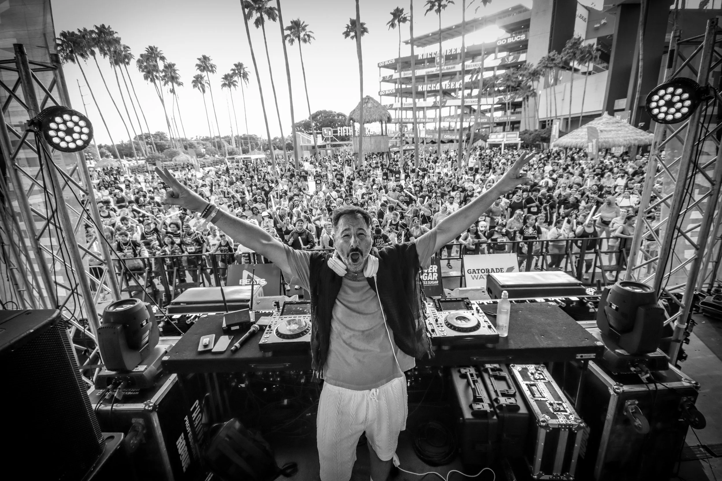 A DJ with arms raised standing behind a DJ booth at an outdoor event, with a large crowd of people in the background, palm trees, and a building.