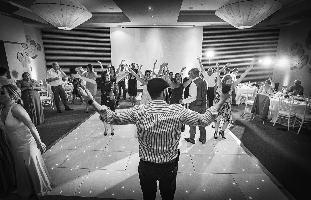People dancing at a party or wedding reception, with a man in the foreground raising his arms on a dance floor, and others dancing and chatting around him in a banquet hall.