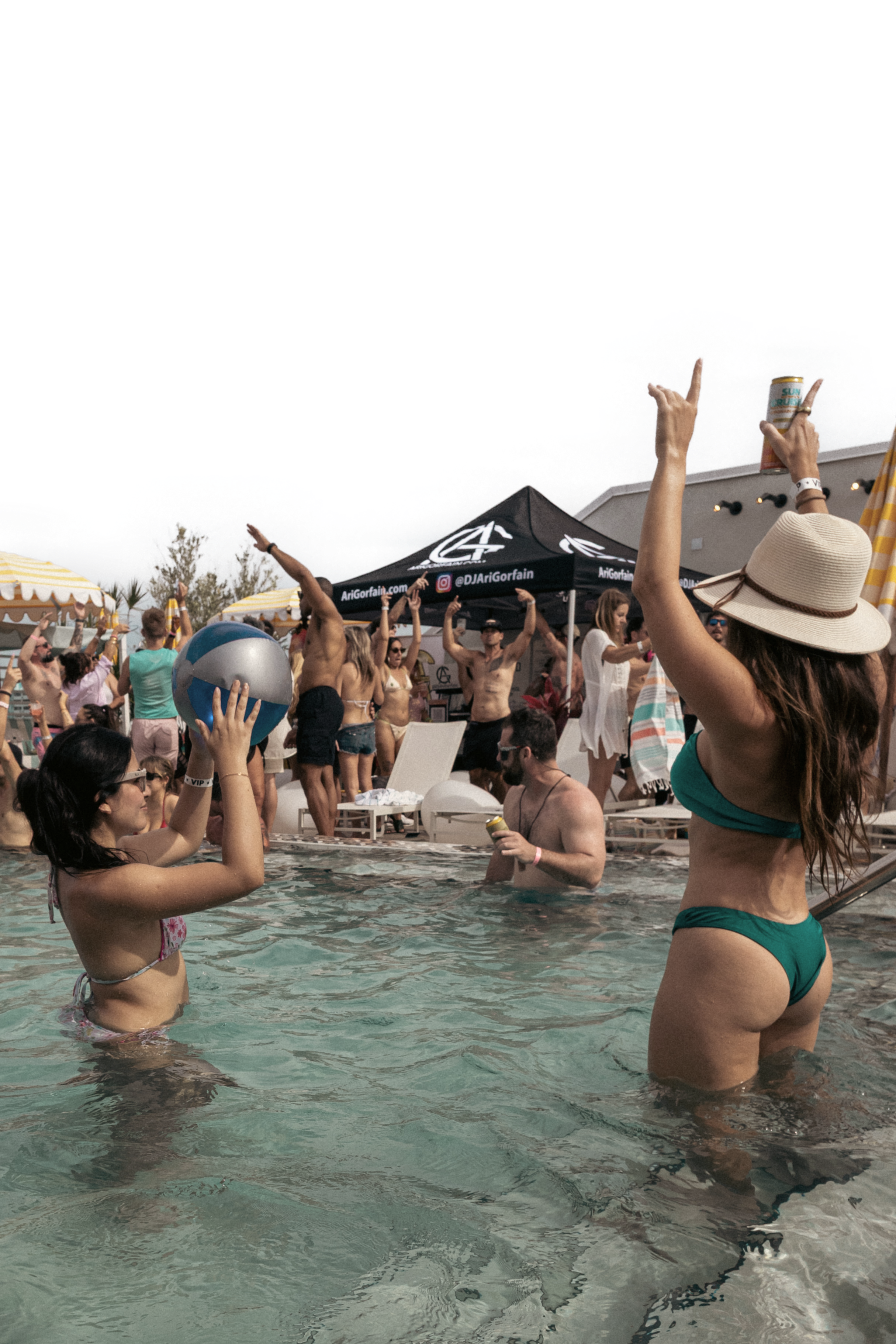 People enjoying a pool party with swimwear, beach balls, and lounging areas under tents, with a building and cloudy sky in the background.