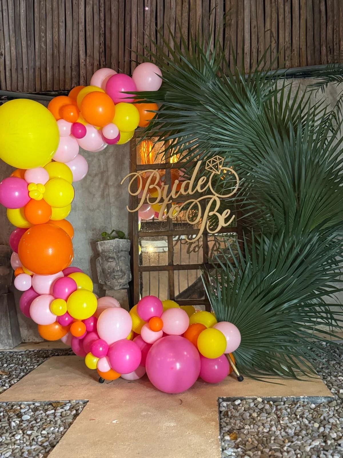 Colorful balloon arch with pink, yellow, orange, and white balloons surrounding a wooden sign that reads "Bride to Be" with greenery and a textured concrete wall in the background.