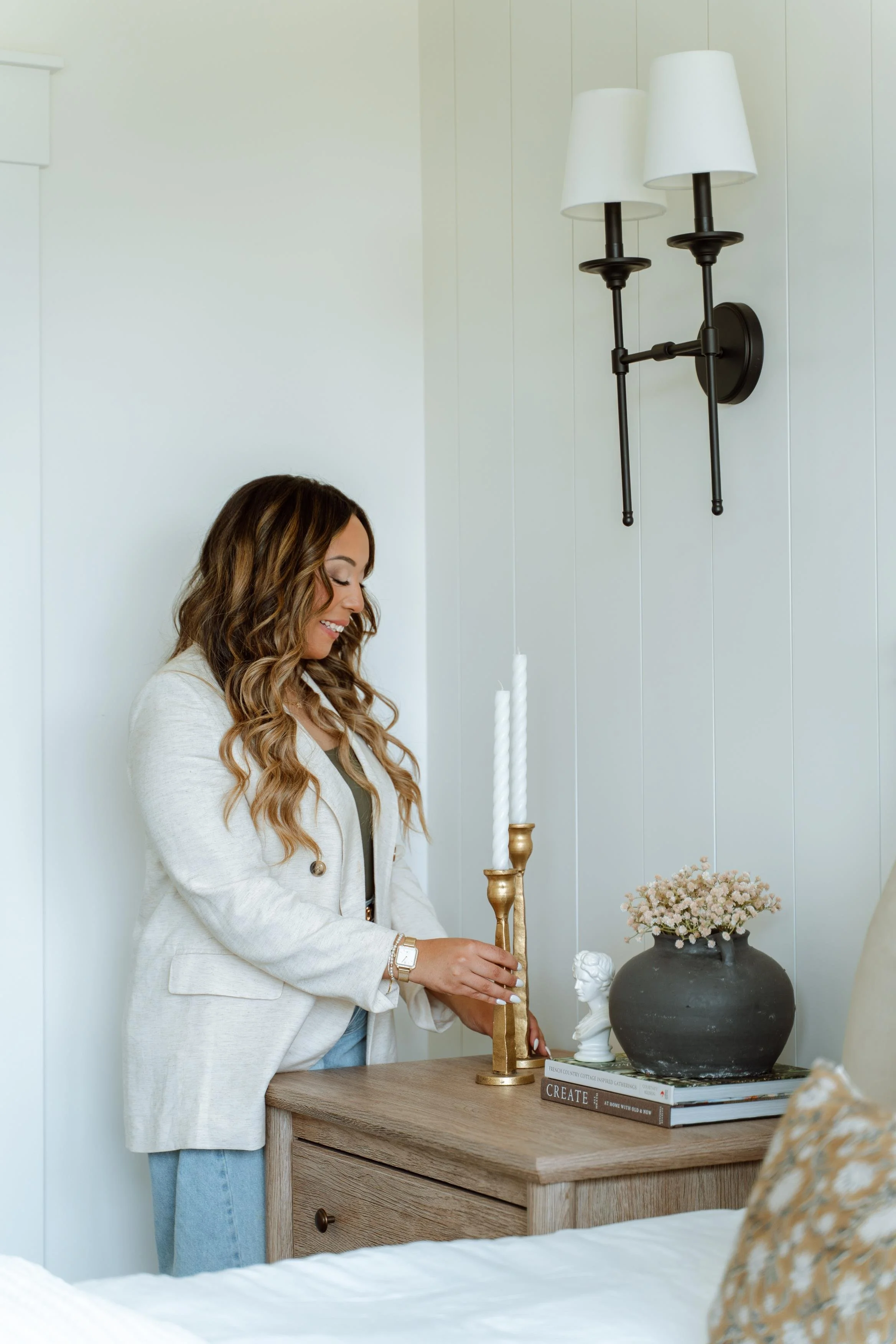 A woman with wavy hair, wearing a light-colored blazer and jeans, arranging candles on a wooden bedside table in a cozy, well-decorated bedroom.