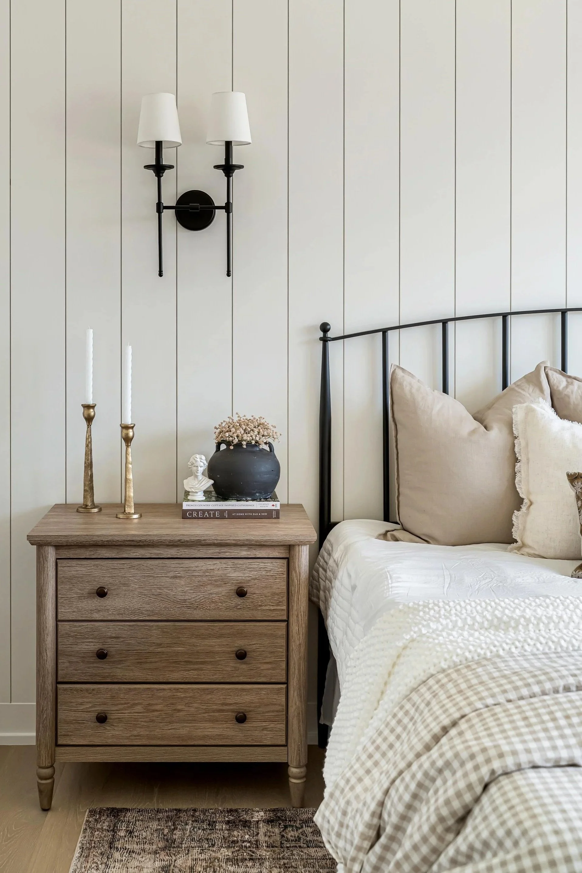 A bedroom corner with a wooden nightstand, a black metal bed frame, a wall sconce with two white lamps, and decorative items including candles, a small bust sculpture, and books.