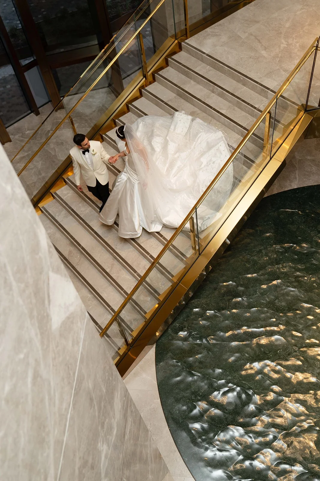 A bride and groom on a staircase, holding hands, in a luxurious indoor setting with water feature.