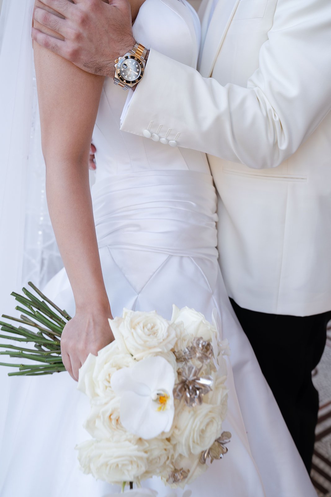 Close-up of a groom in a cream suit holding a bride in a white dress with a bouquet, showing a large gold and silver watch on his wrist.