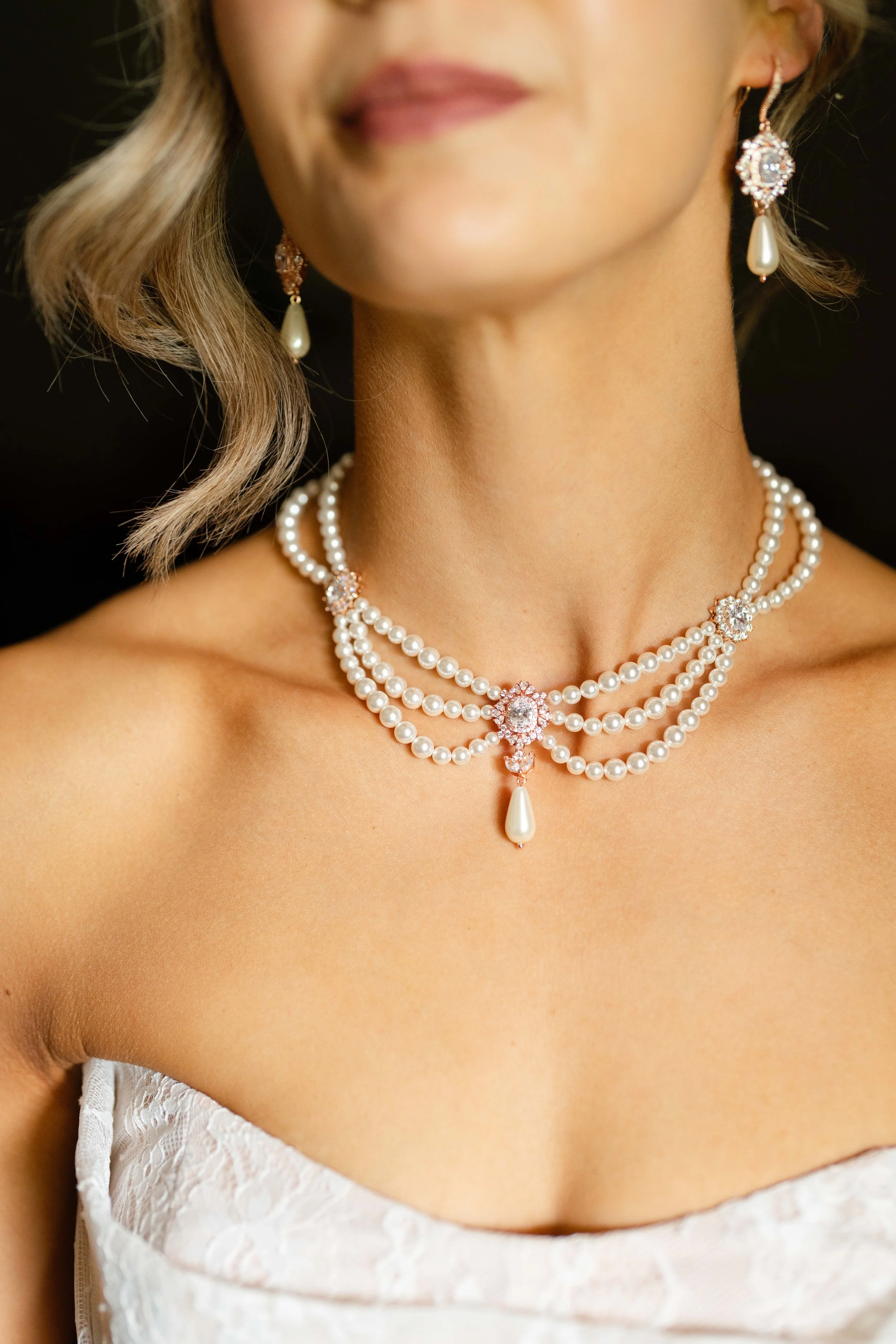 Close-up of a woman wearing pearl jewelry, including a layered pearl necklace with pink gemstone accents and a matching earring with a pearl drop, against a black background.