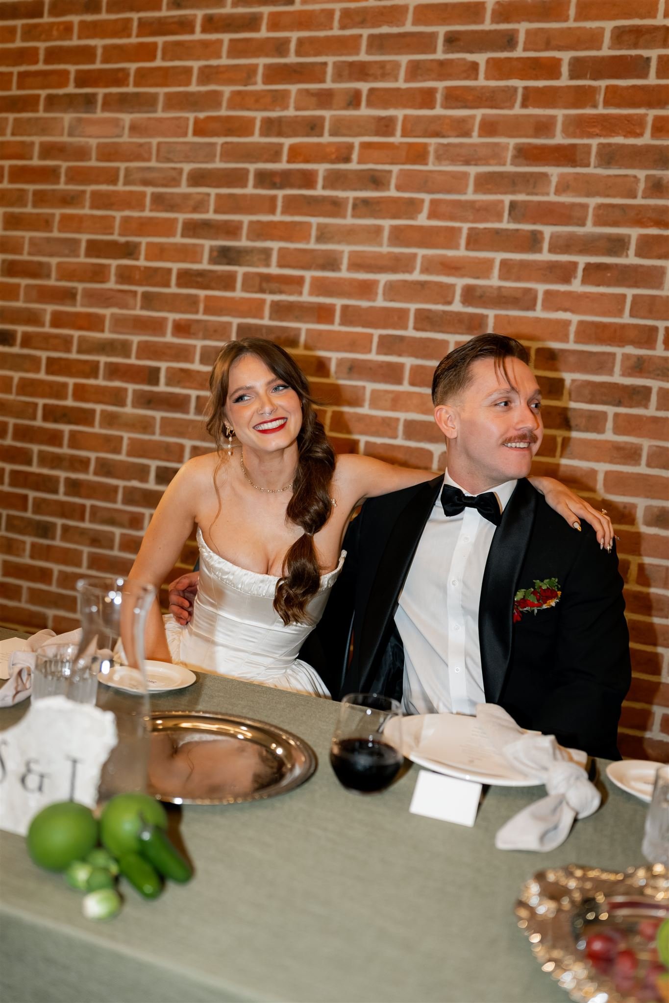 A newlywed couple sitting at a wedding reception table, with the bride wearing a white wedding dress and the groom in a black tuxedo, against a brick wall background.