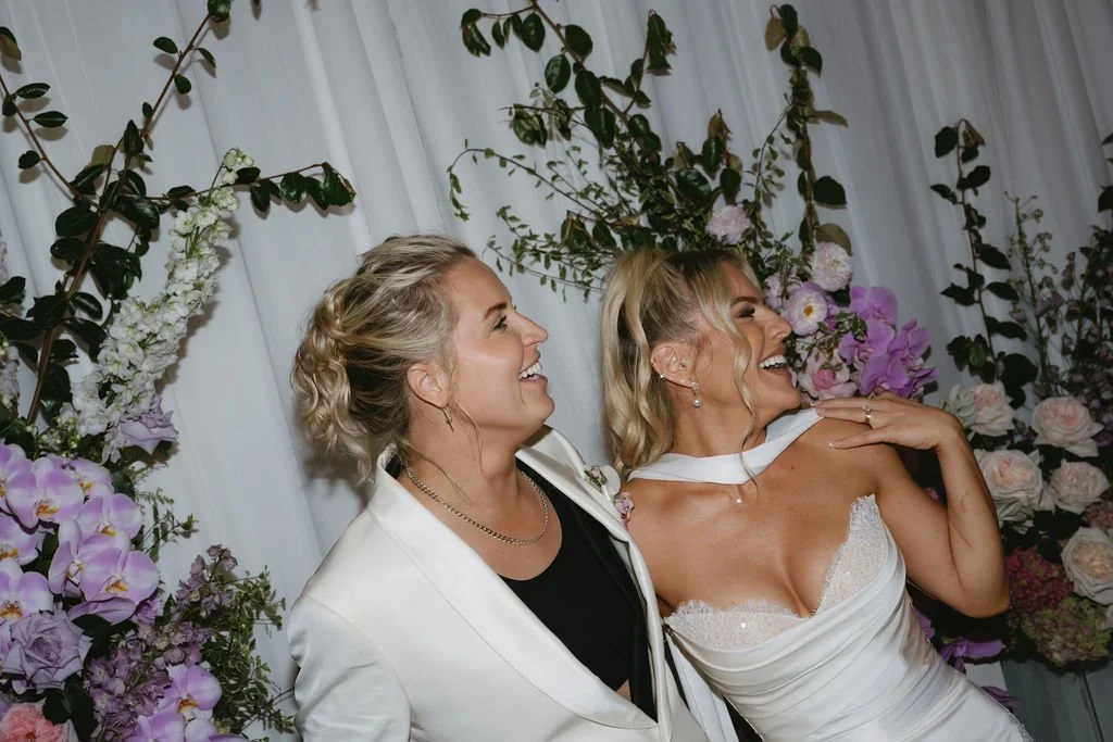 Two women smiling and celebrating at a wedding, standing in front of a floral backdrop with pink and white flowers and greenery.