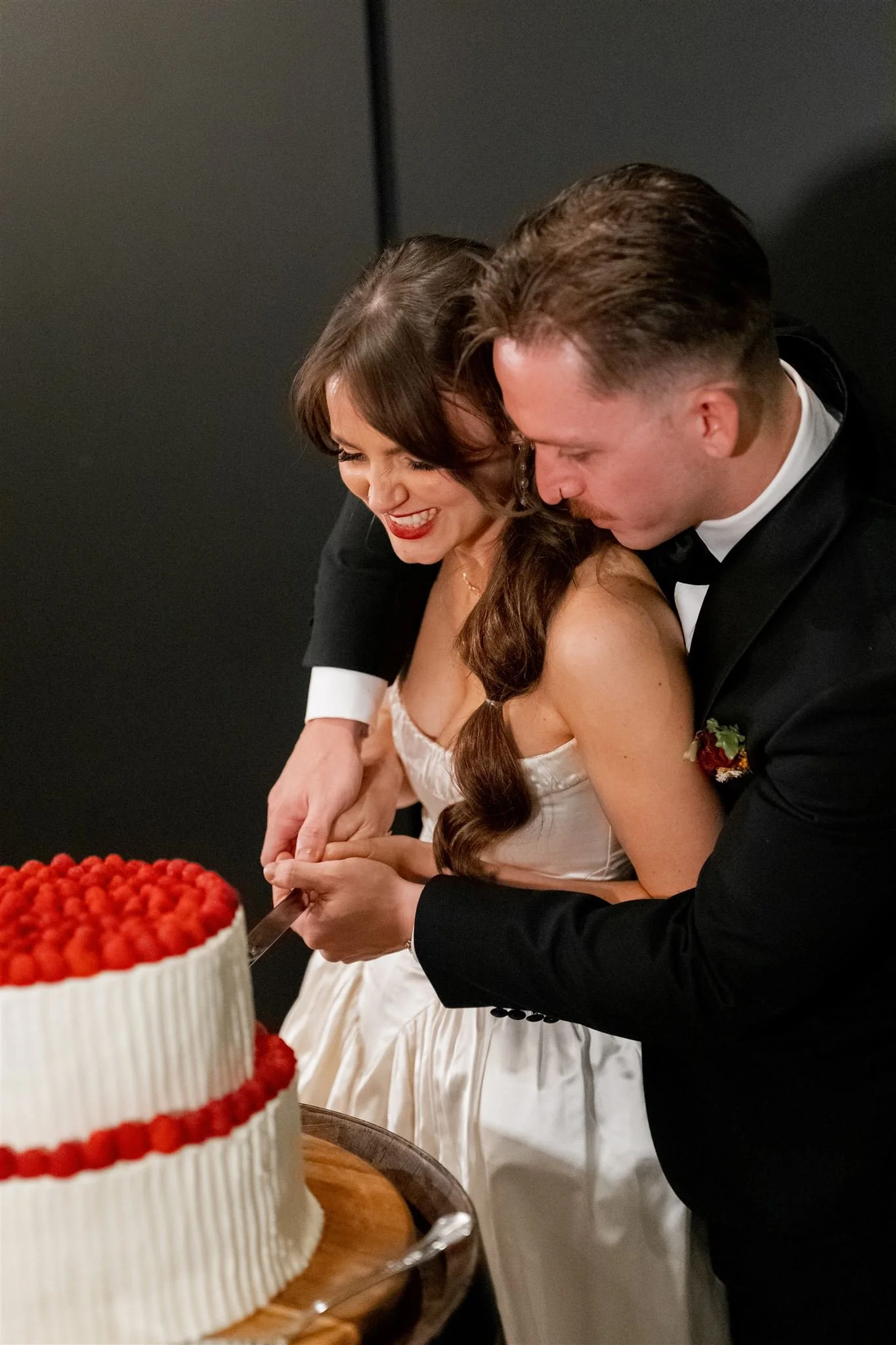 A newlywed couple is cutting a wedding cake together at their reception, smiling and sharing a joyful moment.