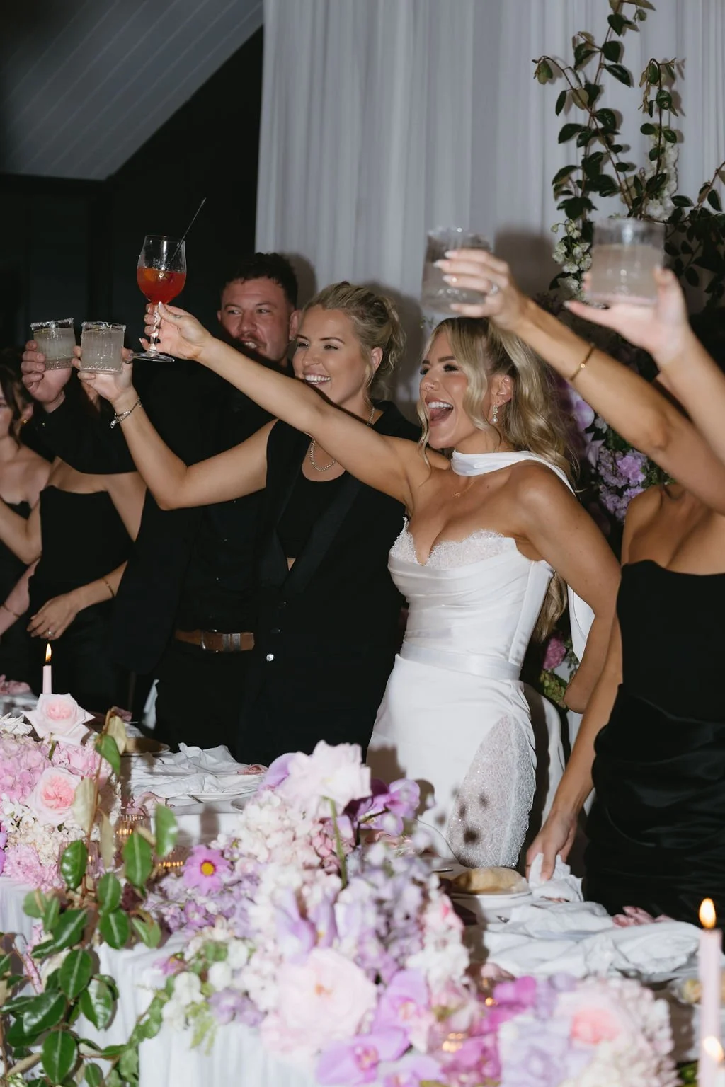 Couples celebrating a wedding toast at a reception with pink flowers and candles on the table.