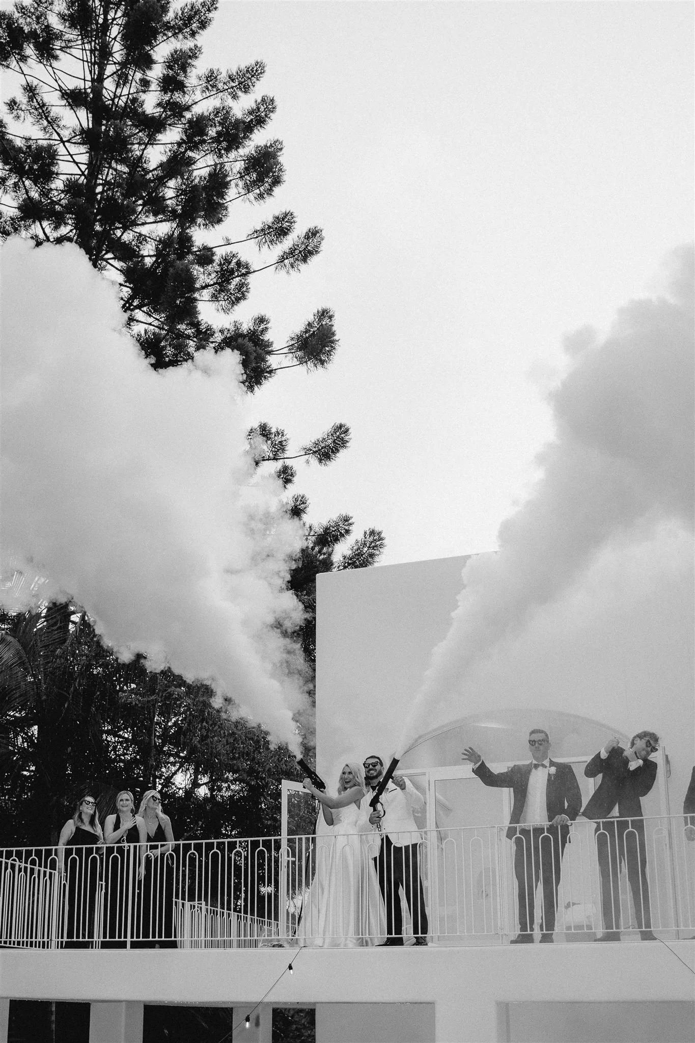 People celebrating on a balcony at a wedding, holding smoke flares, with smoke clouds in the air and tall trees in the background.