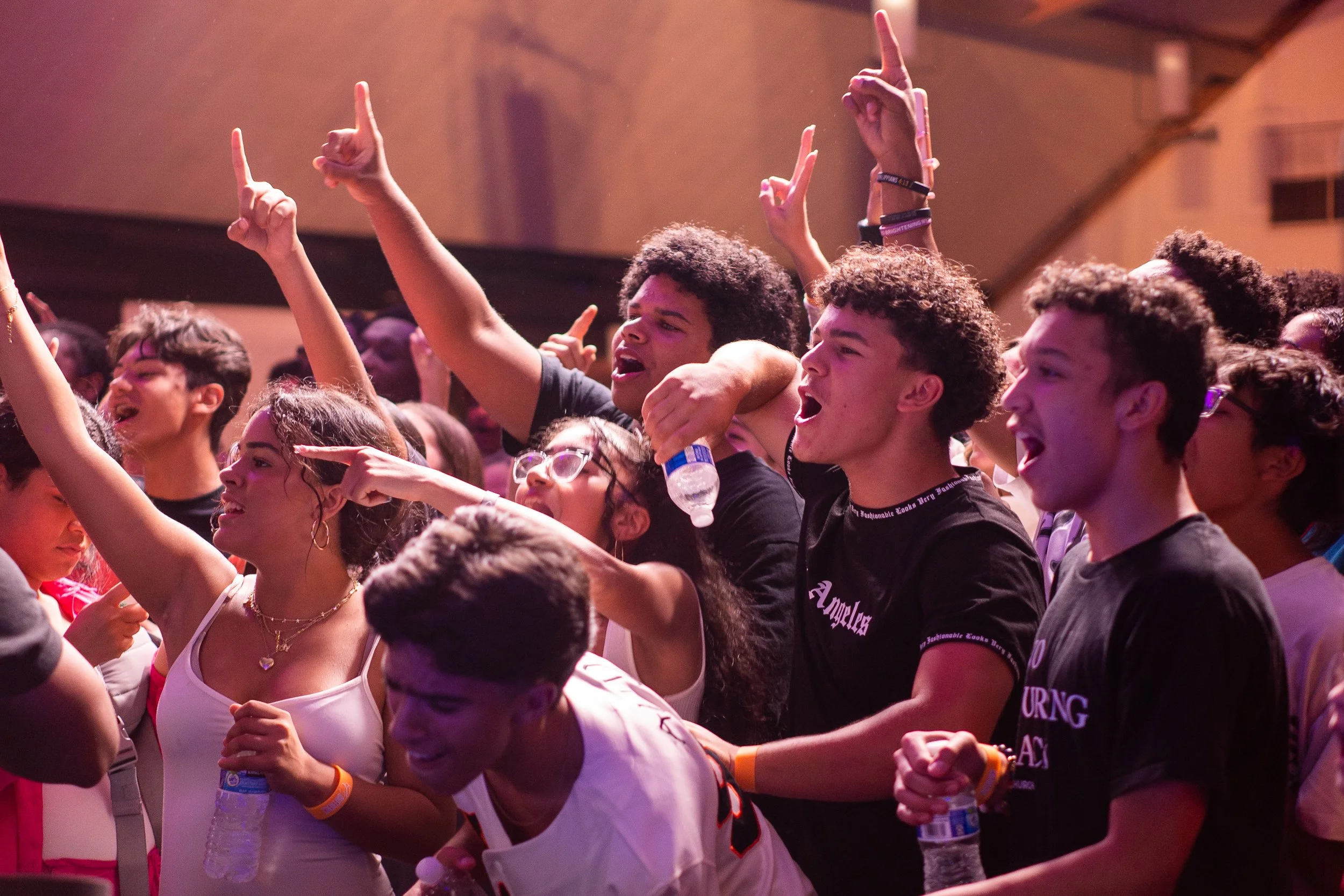 A group of young people at a concert or event, singing, dancing, and enjoying themselves with raised hands and water bottles.
