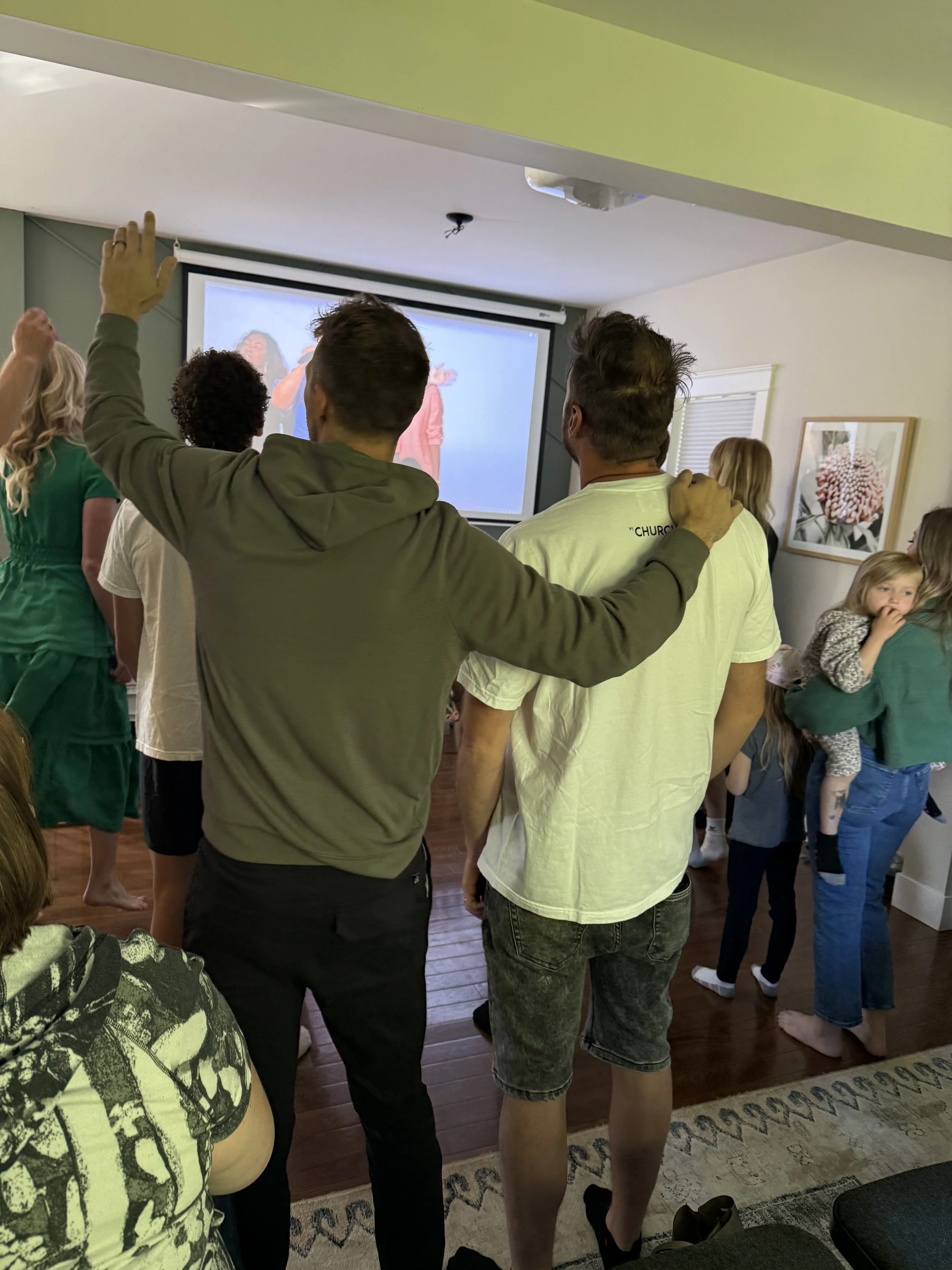 People standing in a living room watching a television, some with their hands raised, including children and adults, with a group gathered near a woman holding a young girl.