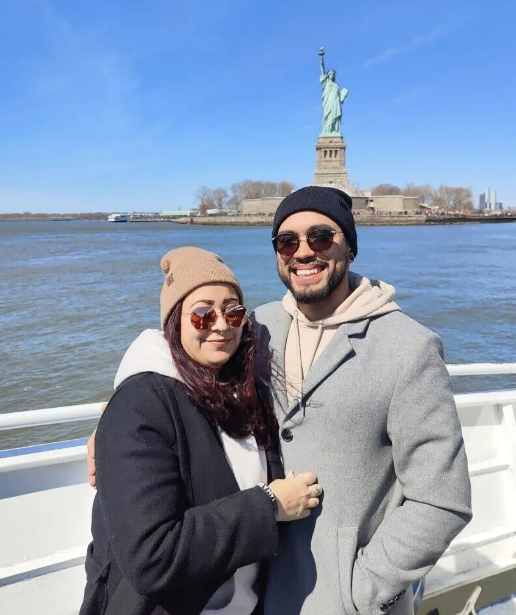 A smiling couple wearing sunglasses and winter clothing on a boat with the Statue of Liberty and New York City skyline in the background.