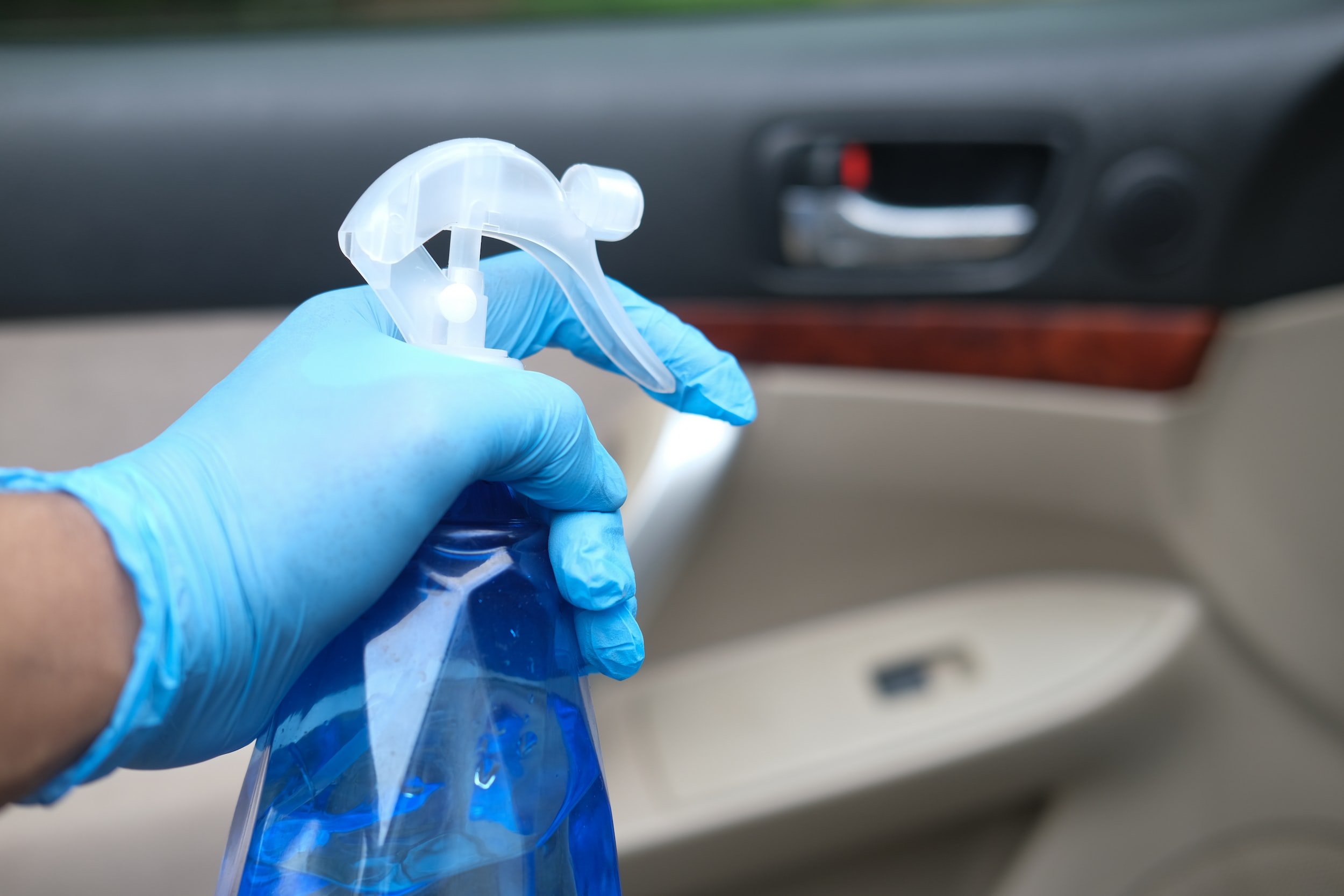 A person wearing blue gloves holds a spray bottle inside a car, with the car door visible in the background.