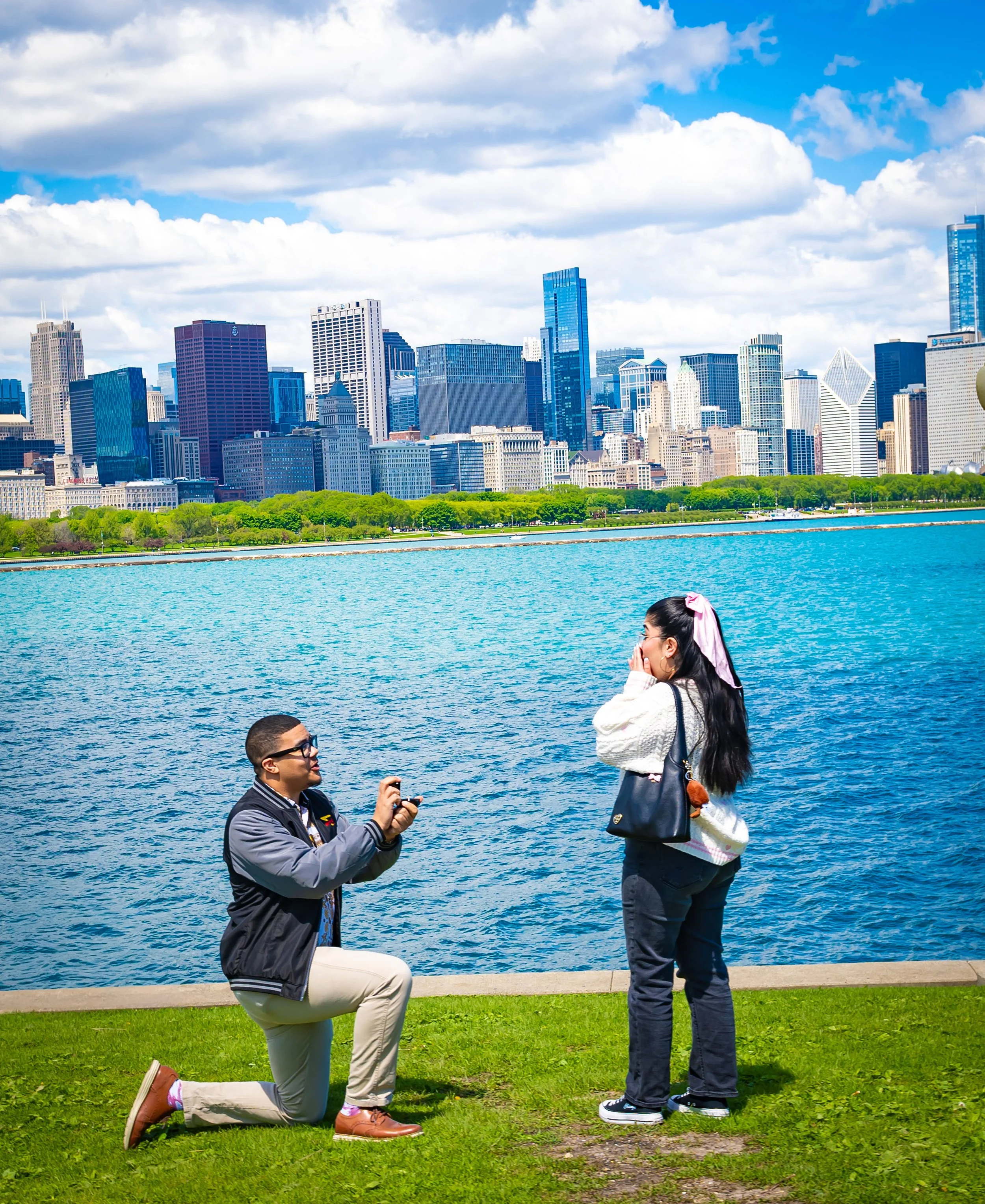 Surprise marriage proposal at Olive Park in Chicago, with a man kneeling and presenting an engagement ring beside Lake Michigan and the Chicago skyline.