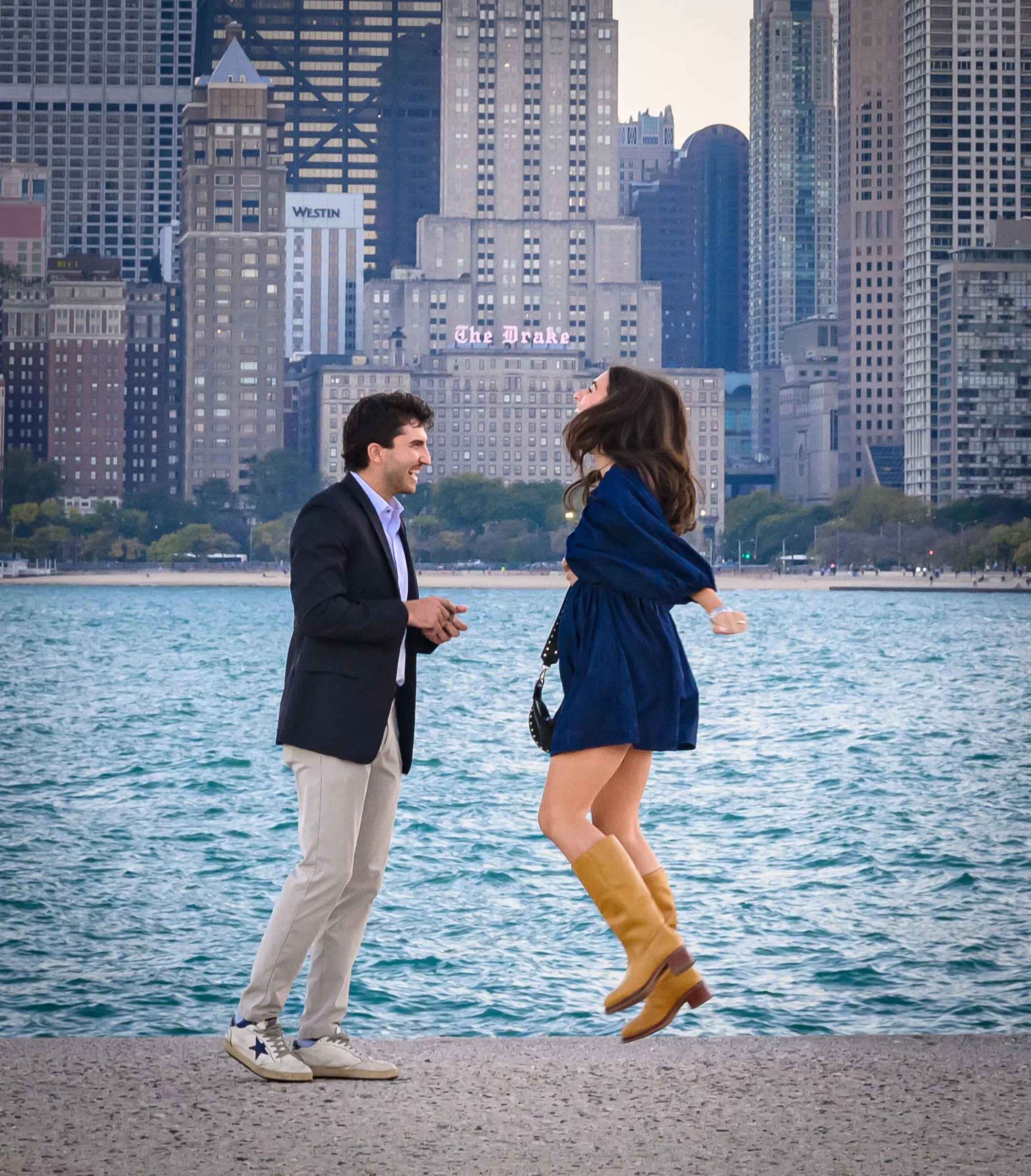 Joyful surprise marriage proposal celebration at North Avenue Beach near Castaways in Chicago’s Lincoln Park. An ecstatic newly engaged woman jumps with excitement beside her fiancé along the Lake Michigan shoreline, with the historic Drake Hotel and