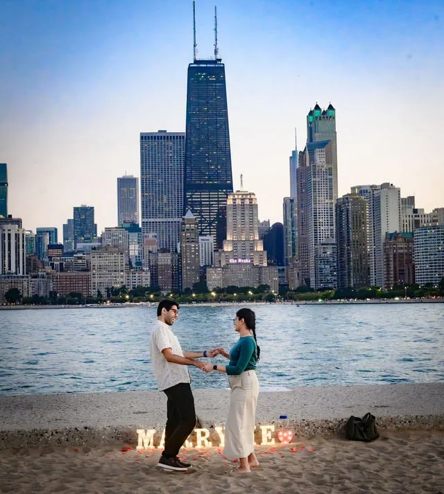 Romantic sunset engagement celebration at North Avenue Beach in Chicago, featuring a newly engaged couple holding hands beside illuminated “MARRY ME” letters and scattered rose petals. Lake Michigan and the iconic Chicago skyline, including the John 