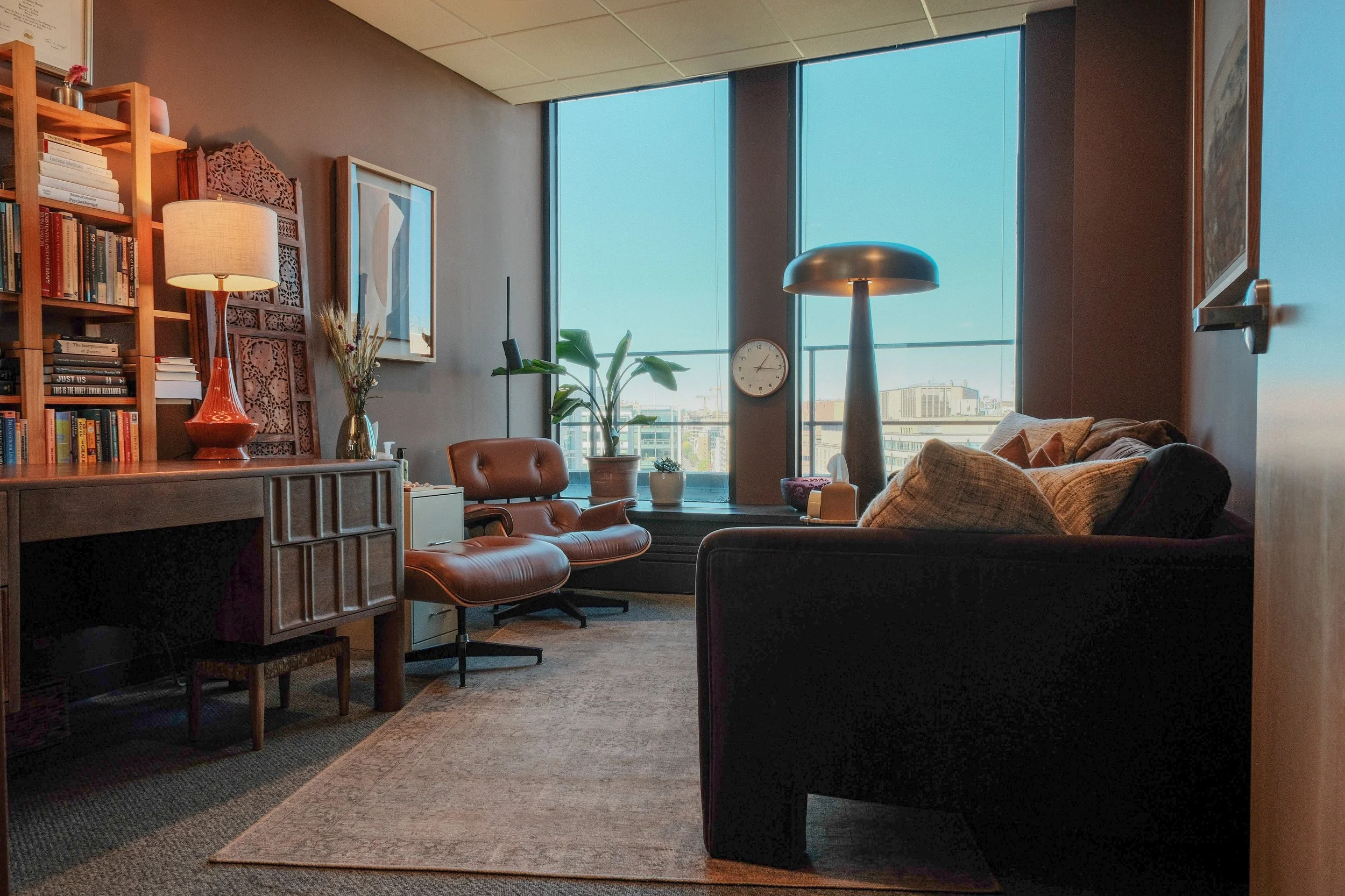 A cozy office with large windows, a brown leather chair, a black sofa with beige cushions, a wooden bookshelf filled with books, a white lamp on a side table, a green plant, a wall clock, and wall art.