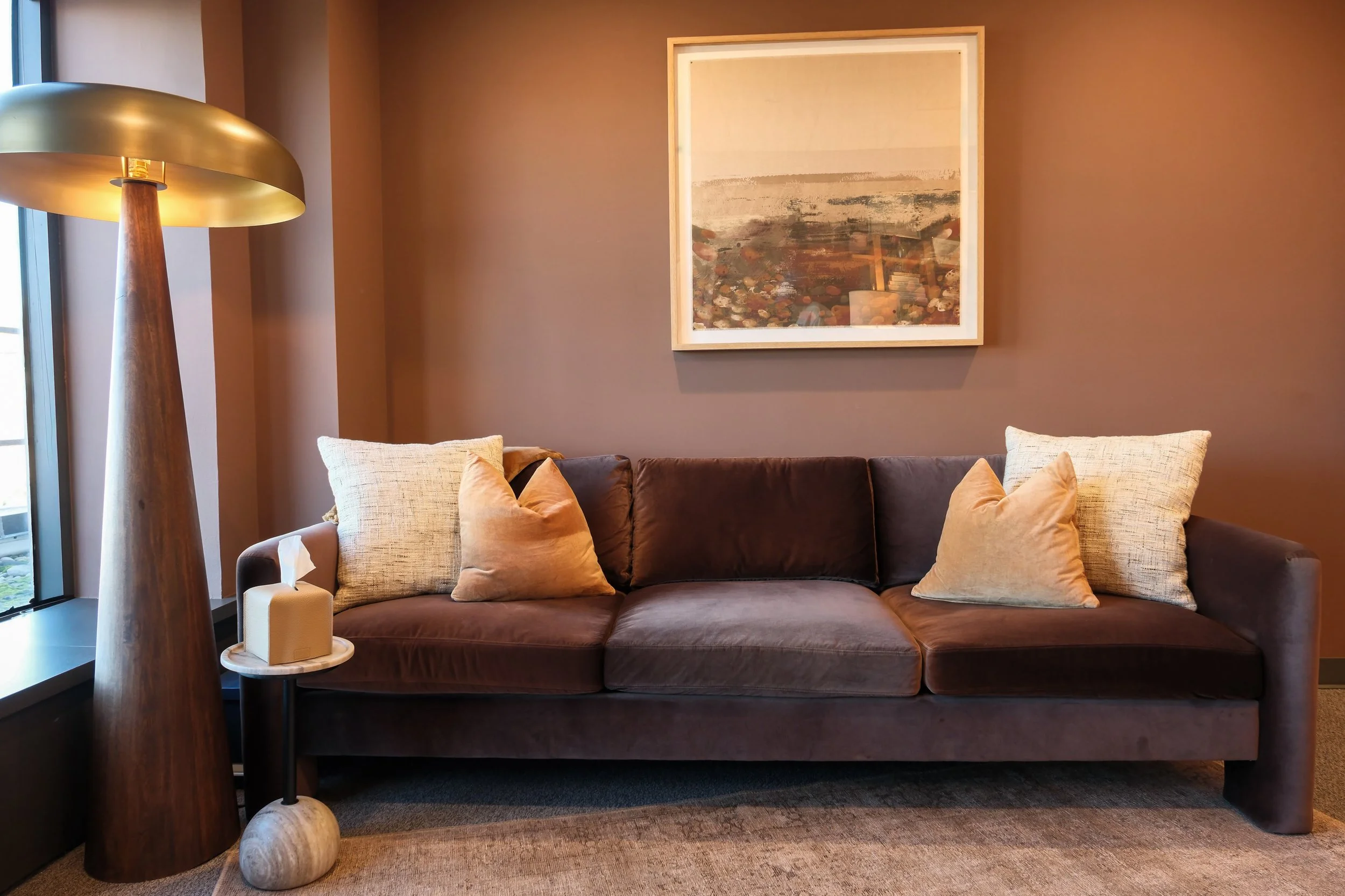 A living room with a brown sofa, multiple beige and orange pillows, a large art piece on the wall, a tall wooden floor lamp, a small round side table with a tissue box and a candle, a window on the left, carpeted flooring, and a warm color palette.