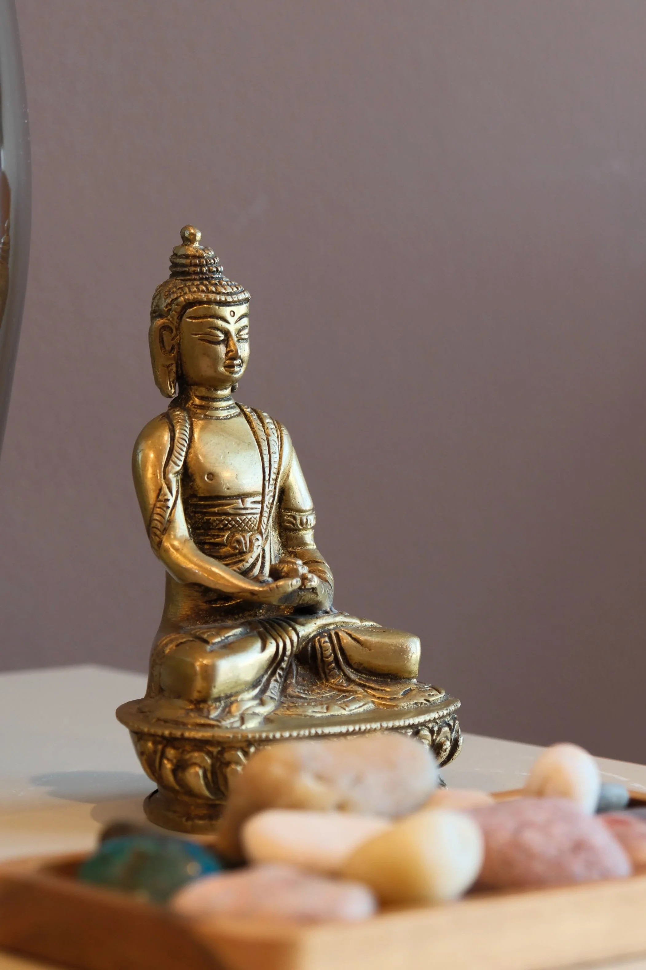 A close-up of a small gold-colored Buddha statue sitting on a decorative base, placed on a table with blurred colorful stones in the foreground.