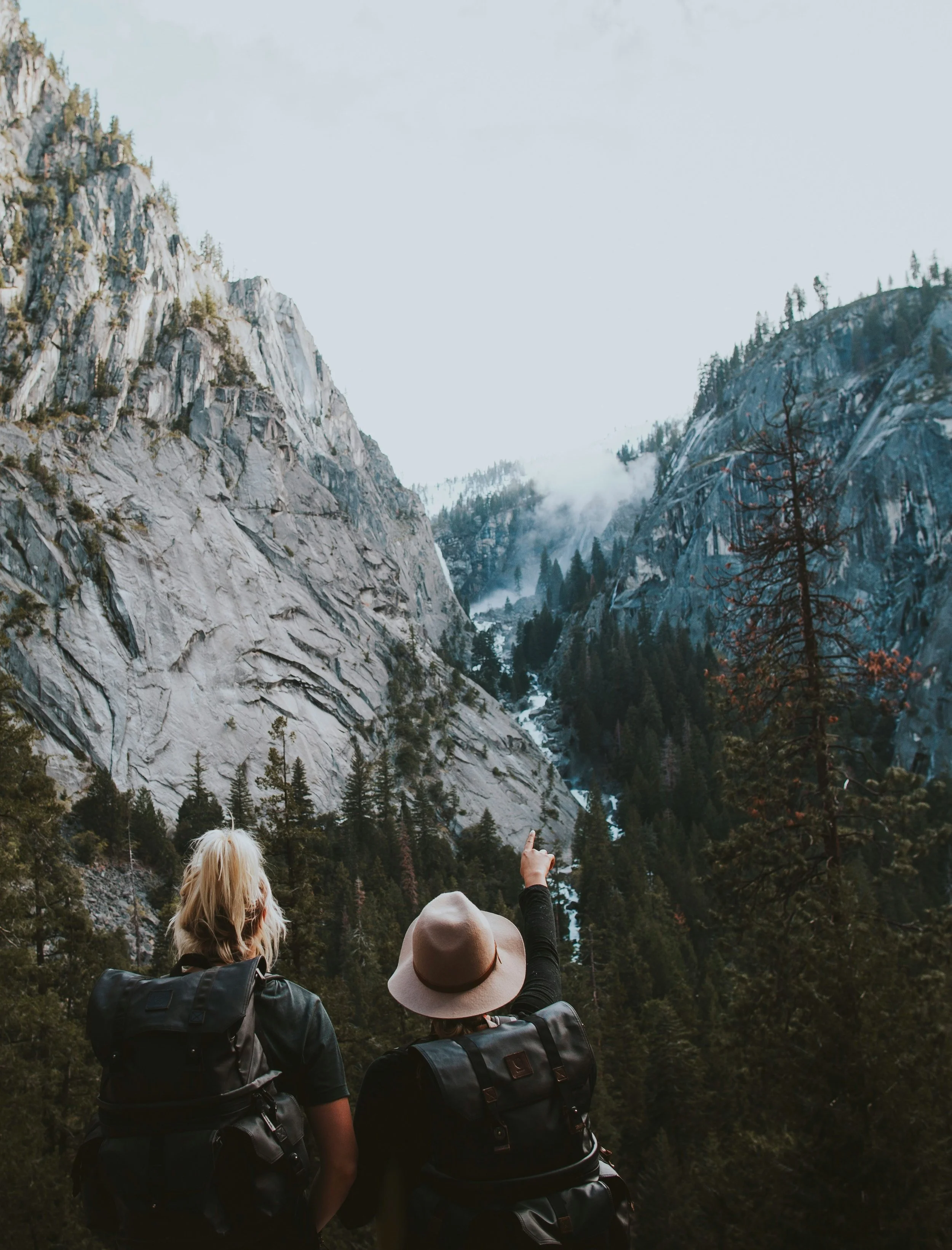 Two hikers with backpacks looking out over a misty mountainous landscape, with one person pointing towards a distant peak