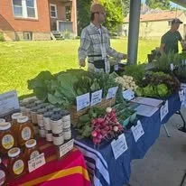 A man standing behind a table full of green vegetables and radishes at the LoveCarrick pavilion. Another table shows honey and rootbeer. 