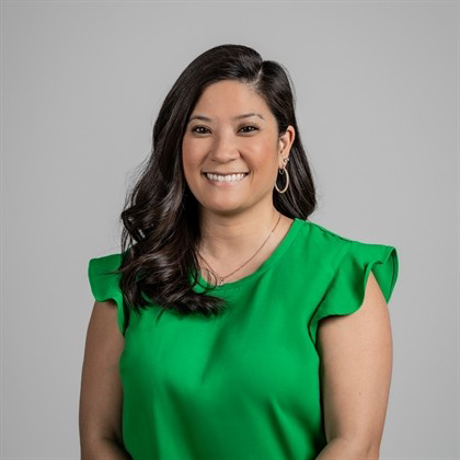 A woman with long dark wavy hair wearing a green top and hoop earrings, smiling against a gray background.