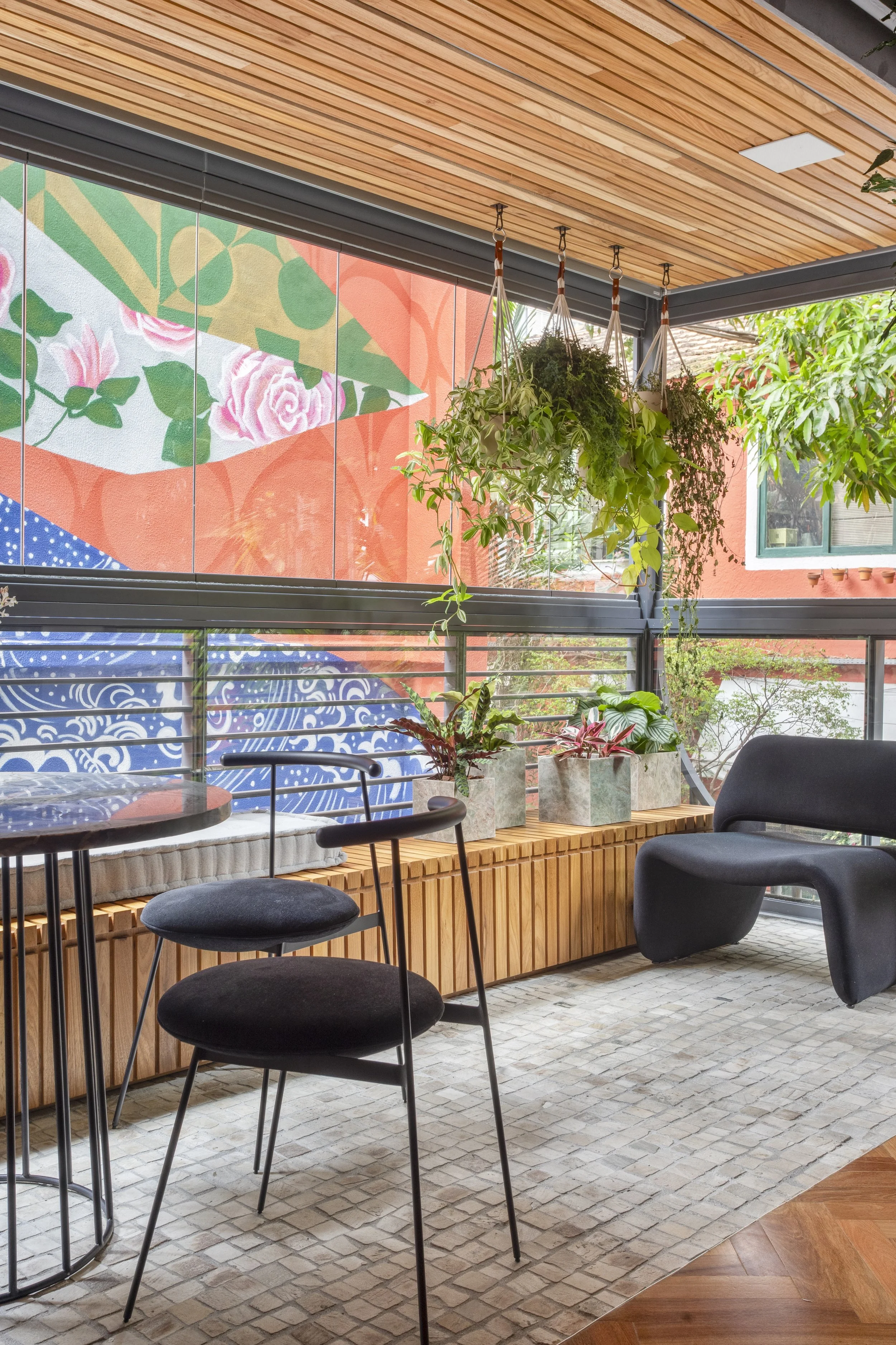 Modern indoor seating area with black chairs and a round table. Hanging plants are suspended from a wooden slatted ceiling. In the background, a large colorful mural is visible through glass windows.