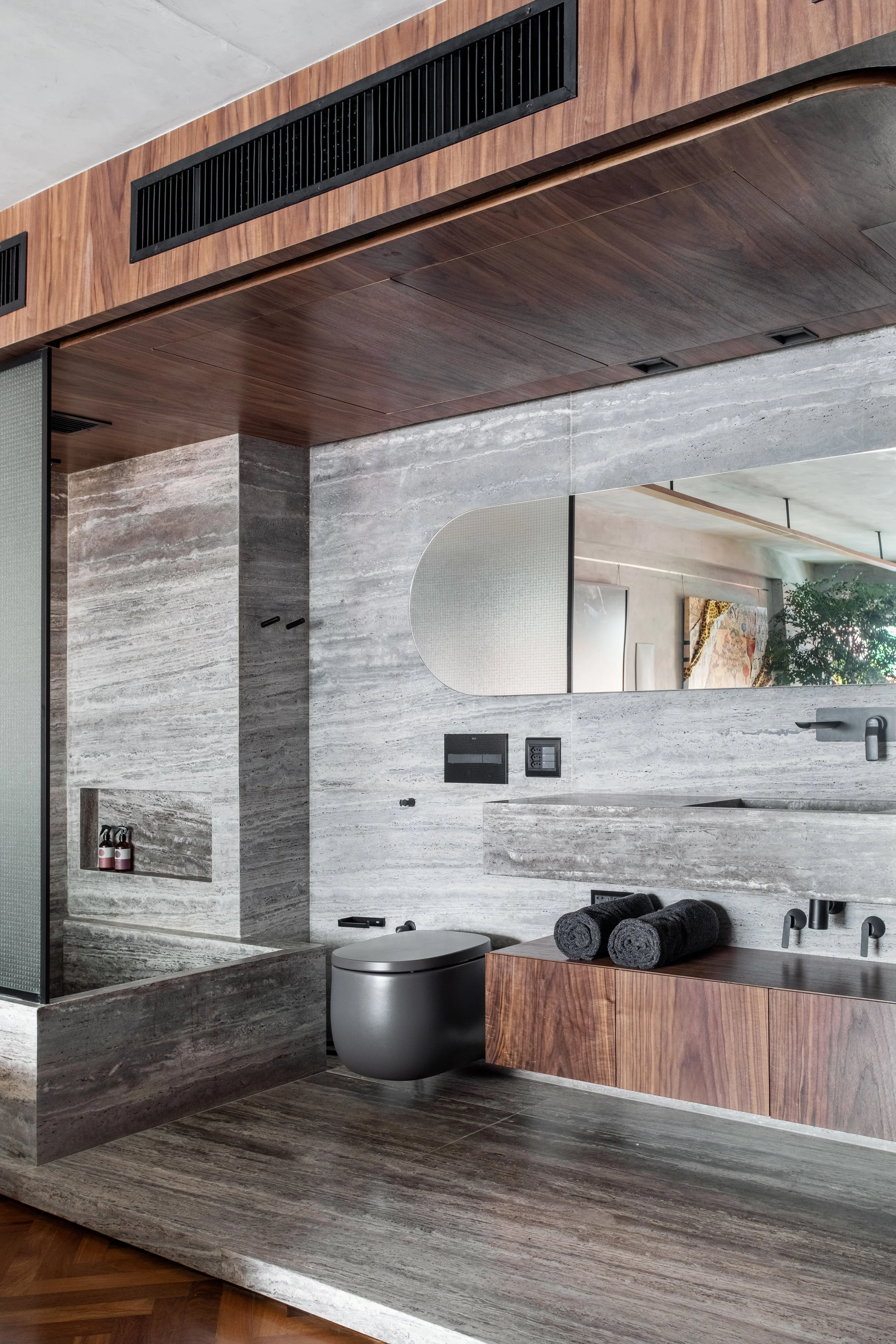 Modern bathroom with gray stone walls, wood accents, an oval mirror, black toilet, and built-in shelves.