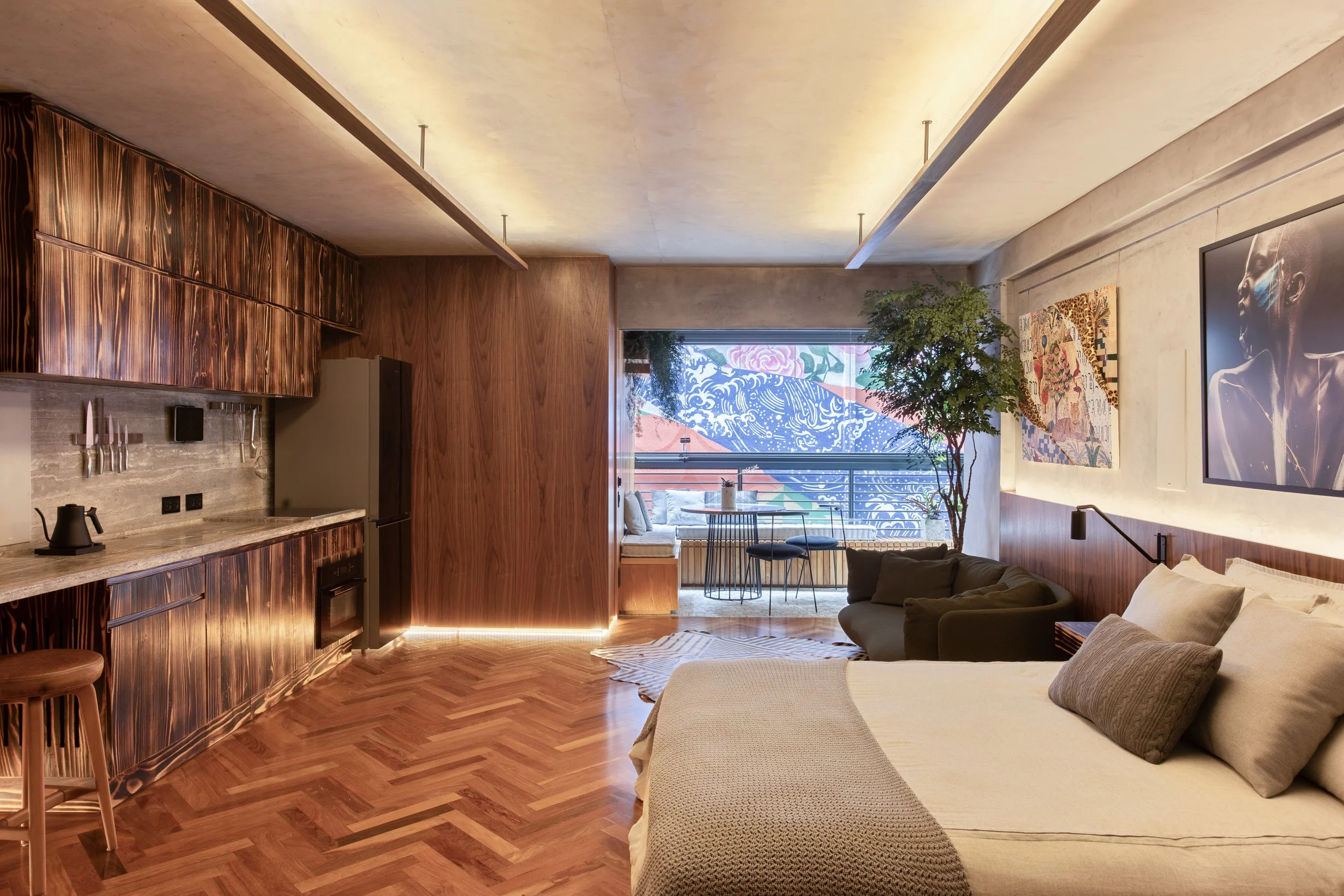 Modern studio apartment with wood accents, featuring a bed, small kitchen area, and a seating space by a large window with colorful mural outside. The room has parquet flooring, a cozy chair, and decorative wall art.