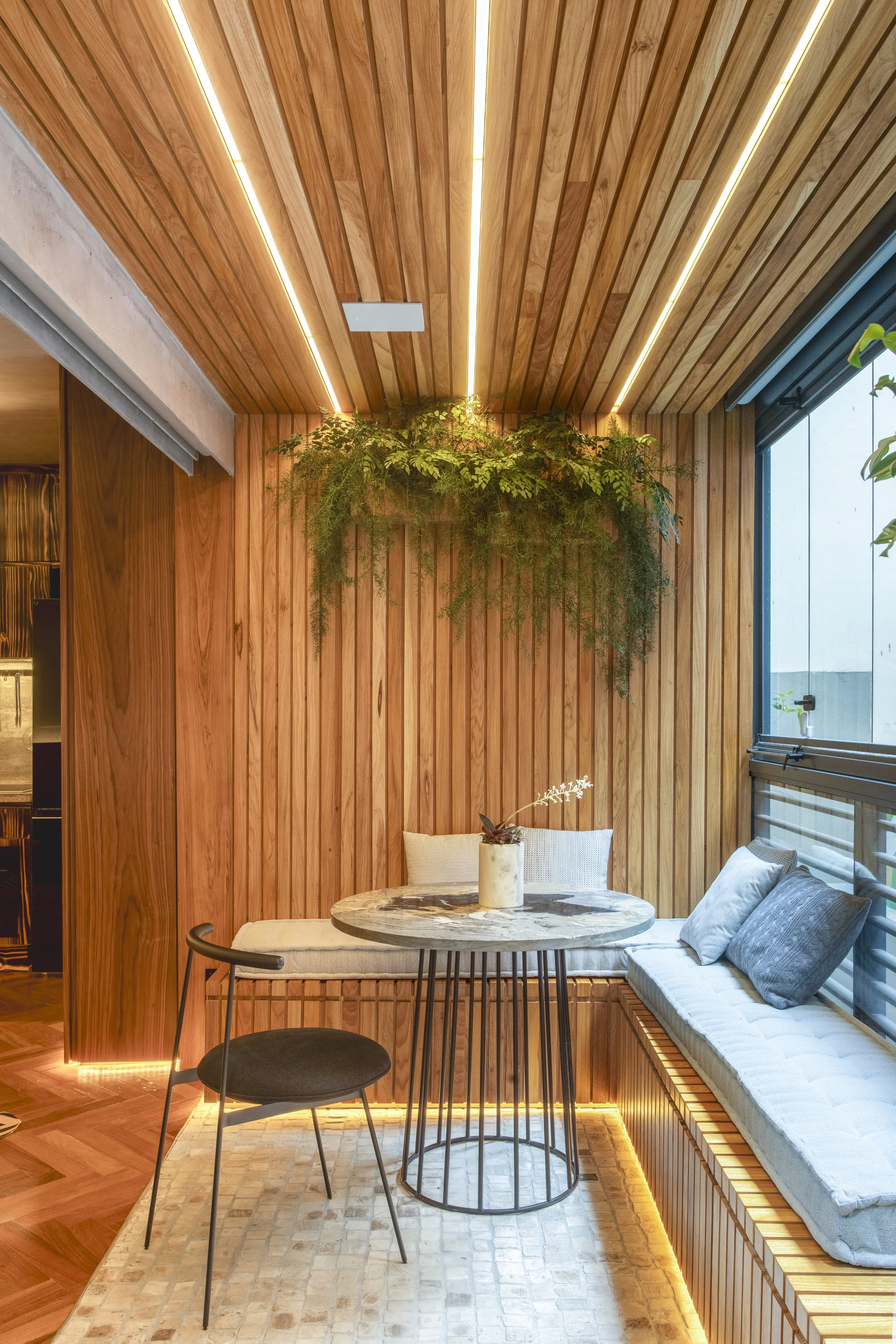 Modern cozy corner with wooden slat ceiling and walls, featuring a round table, black chair, and built-in bench seating with cushions. Greenery hangs from the ceiling.