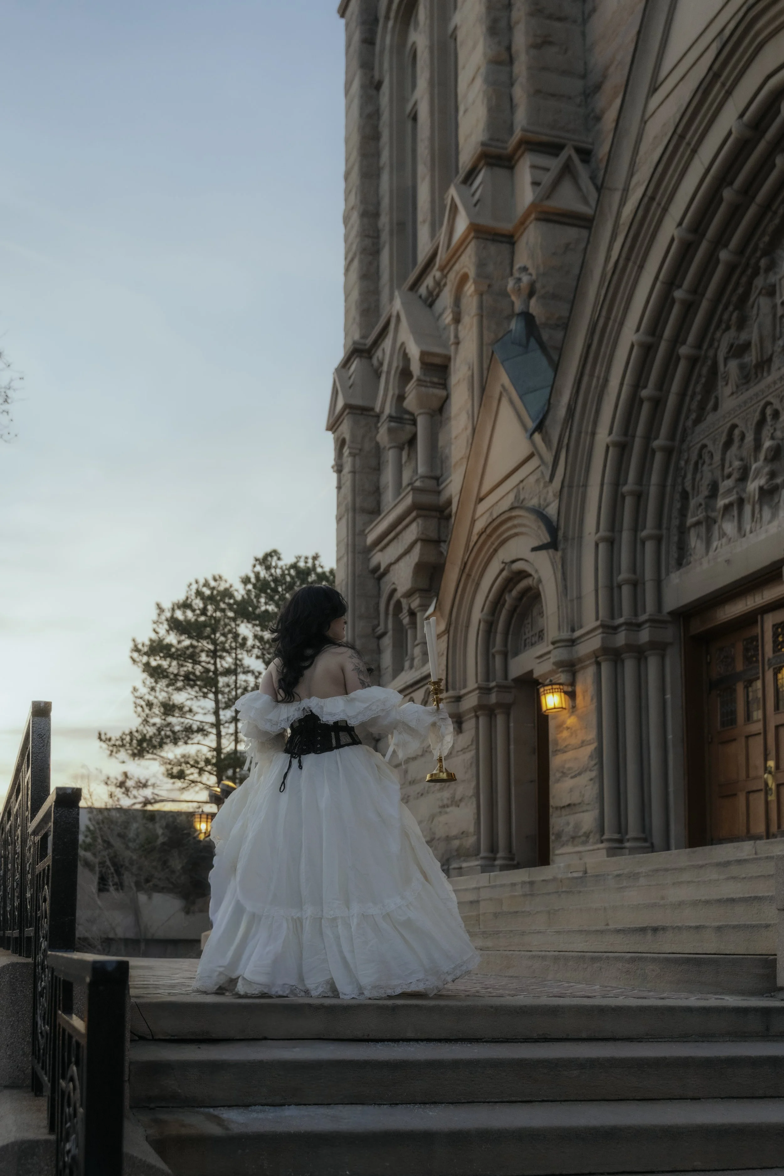 A woman in a vintage, off-shoulder white gown holding a candlestick stands on the steps of a stone church at dusk, facing away from the camera.