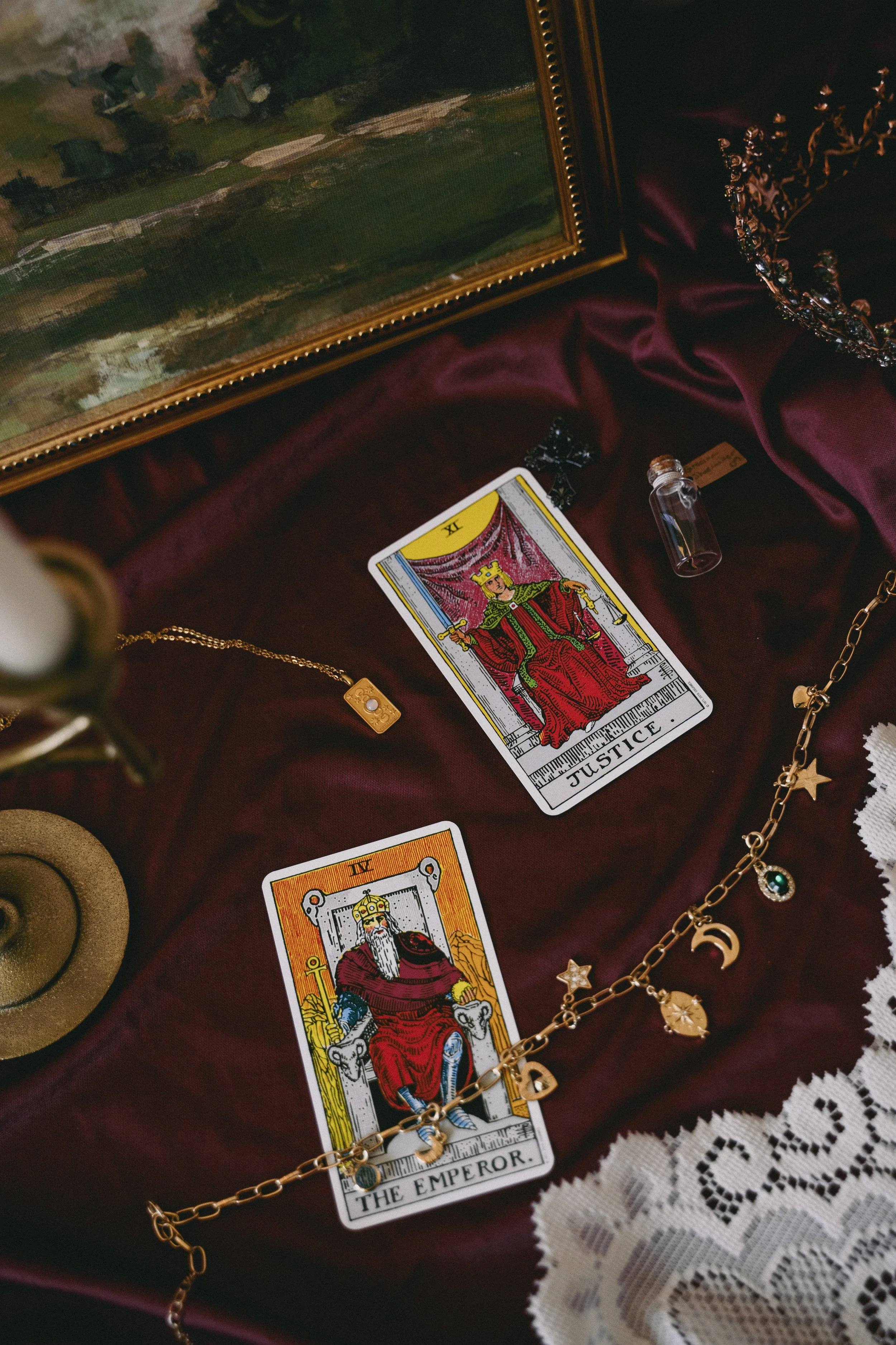 Two tarot cards, Justice and The Emperor, are laid out on a deep red cloth. Surrounding them are jewelry items, including a gold necklace with star and moon charms, a small glass vial, and a black cross pendant. In the background, part of a framed painting and lace fabric are visible.