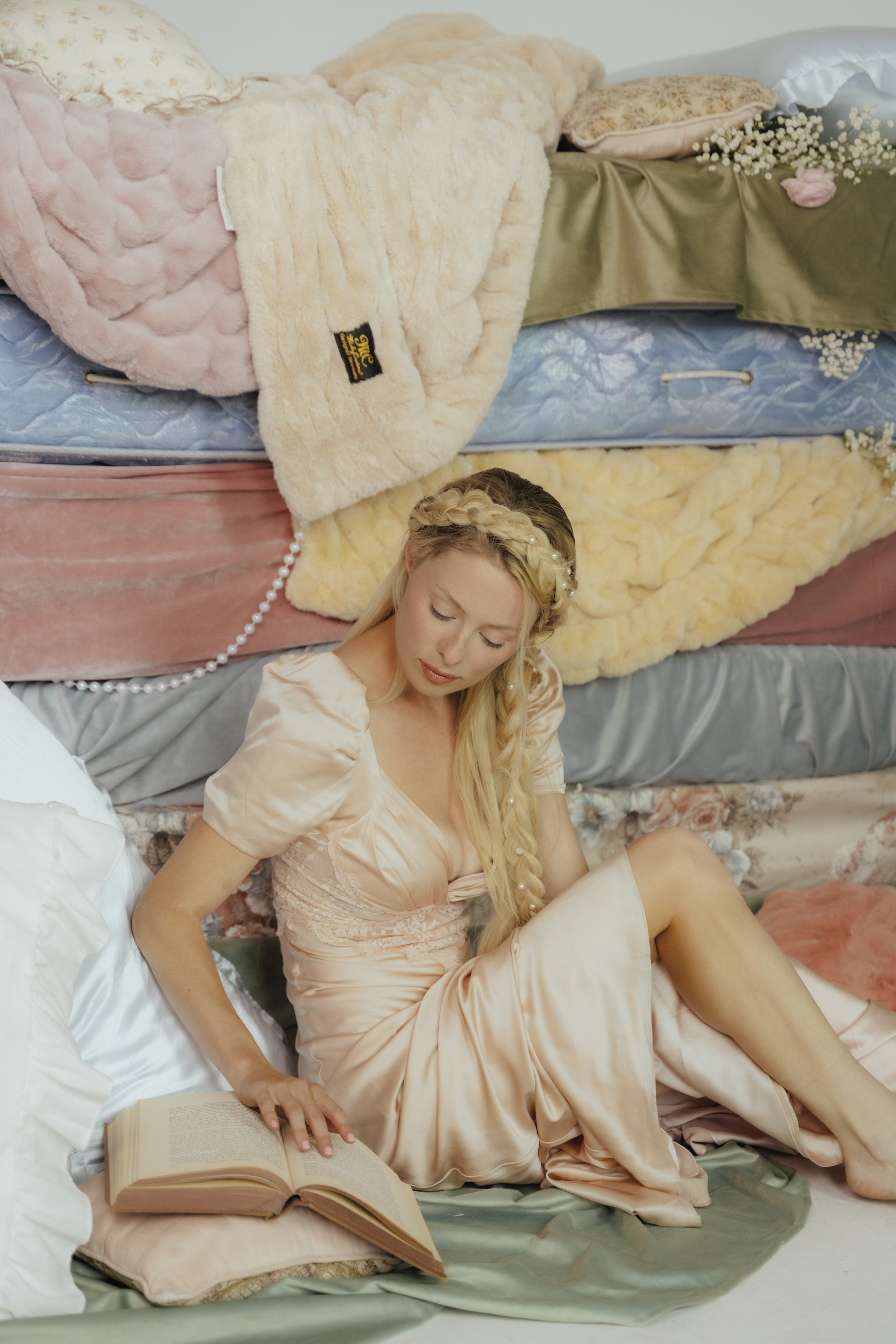 Young woman with long blonde hair braided, wearing a satin dress, lying on a bed surrounded by pillows. She is reading a book with her hand on the open pages, and there is an open book next to her.