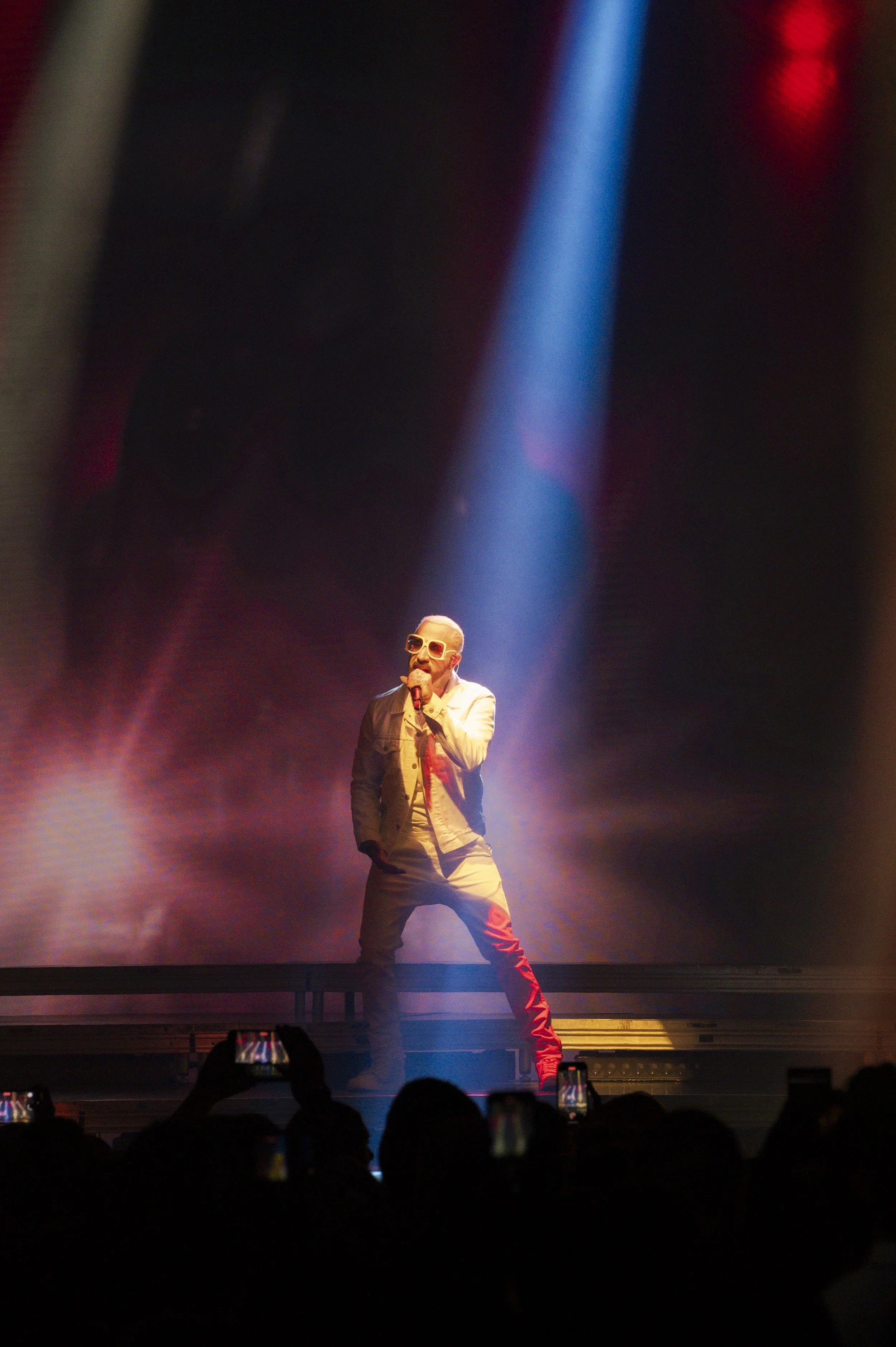 Performer singing on stage under blue spotlight, holding microphone, wearing sunglasses, and casual outfit, with audience taking photos.