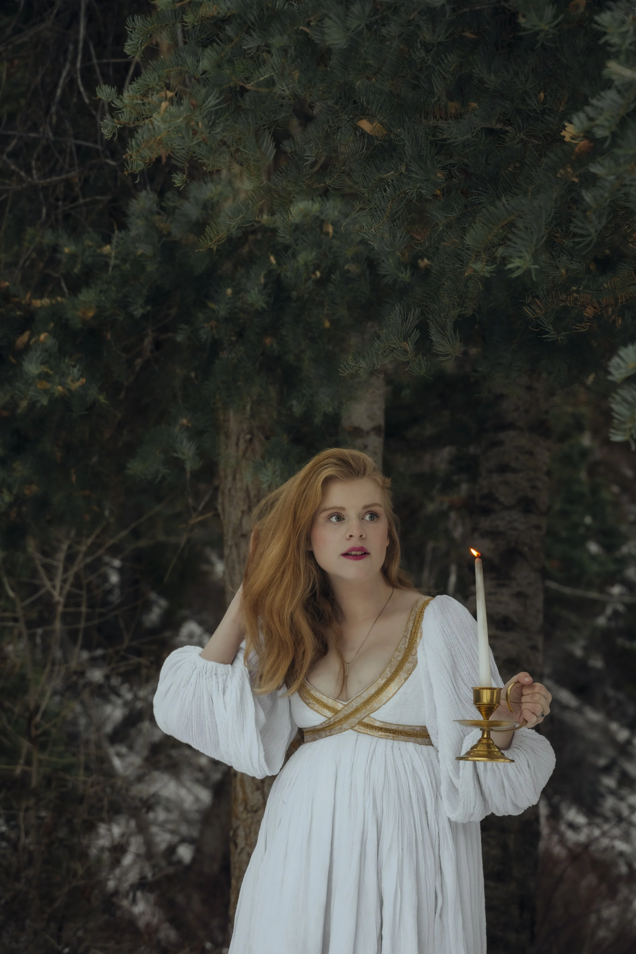 A woman with red hair wearing a white dress with gold embroidery holding a brass candlestick with a lit white candle in a forest setting.