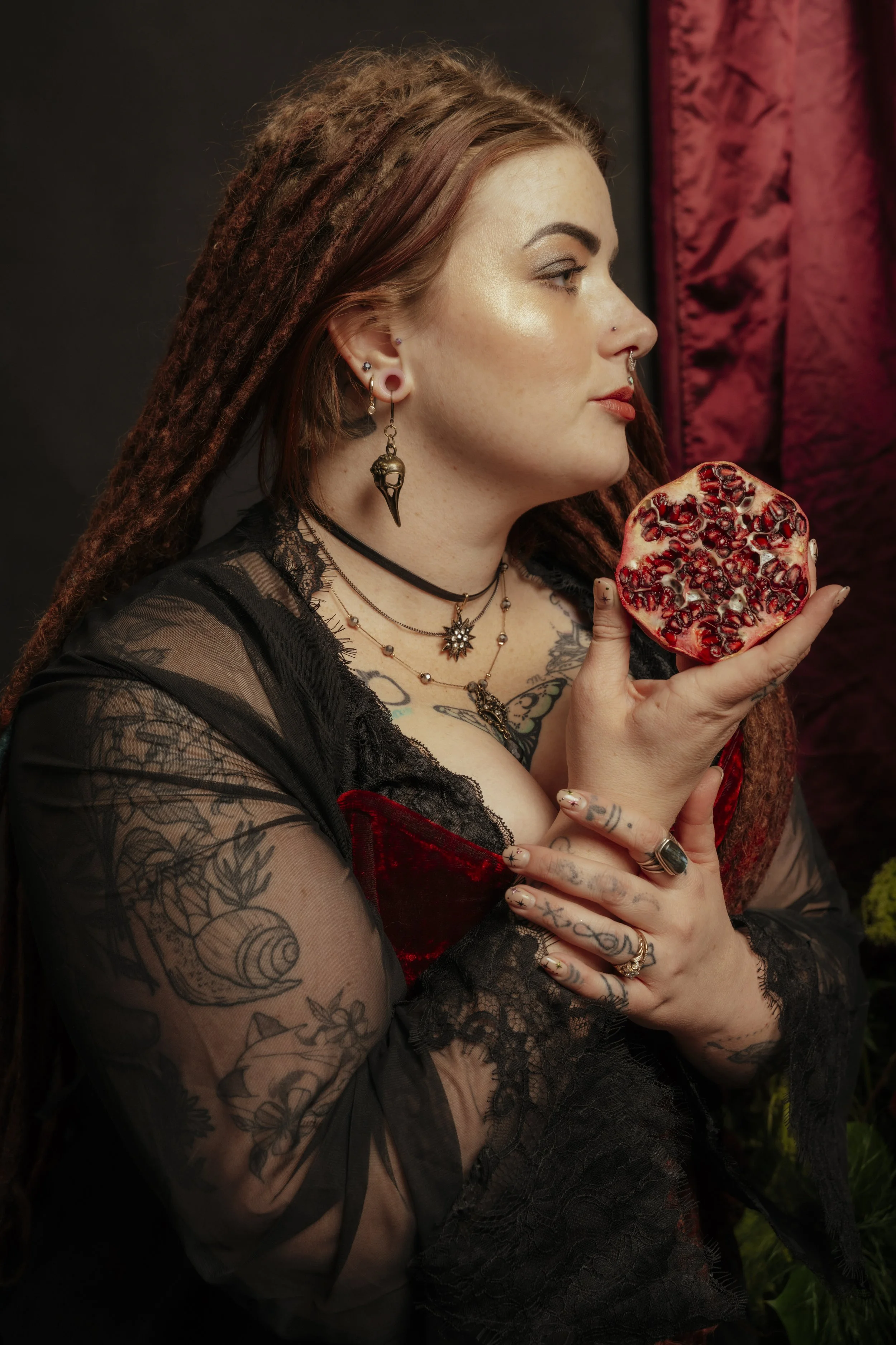 A woman with tattooed arms and multiple piercings, dressed in Gothic style, holds a pomegranate half close to her face, looking to the side. She has long, reddish-brown dreadlocks, and is wearing layered necklaces and earrings. She is in a dark setti