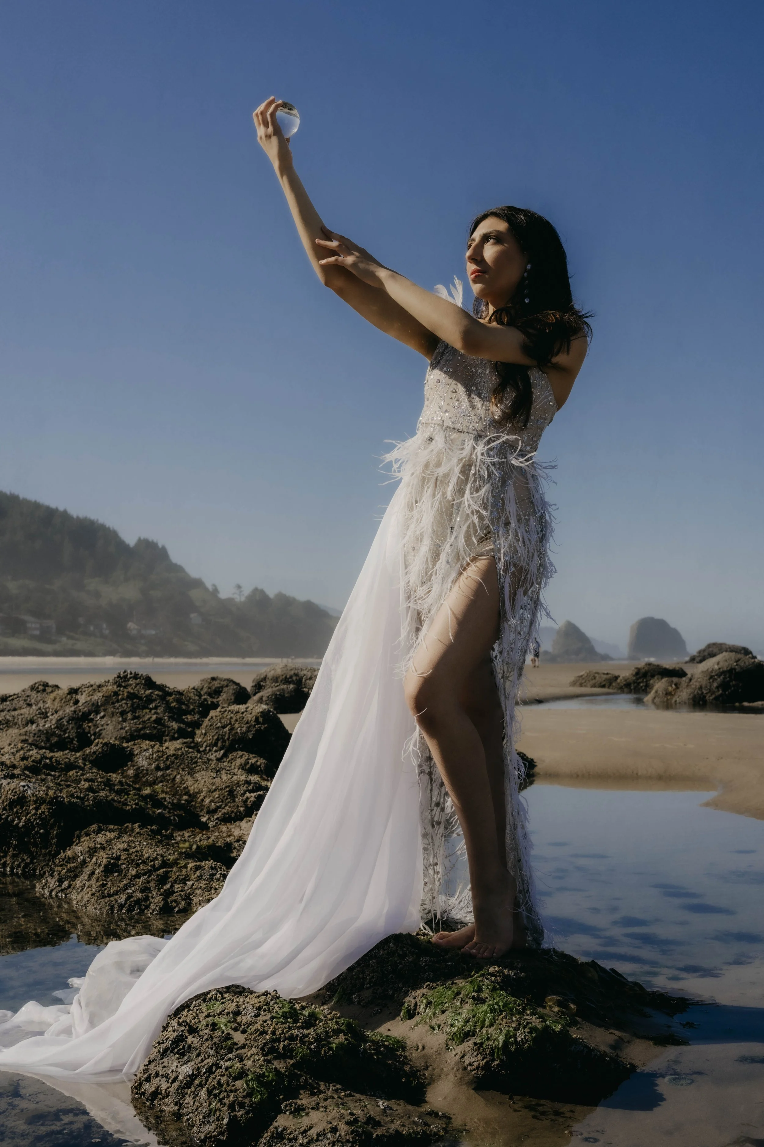A woman in a long, flowing white dress standing on rocks at the beach, holding a crystal ball overhead, with a clear blue sky and distant rocky formations in the background.