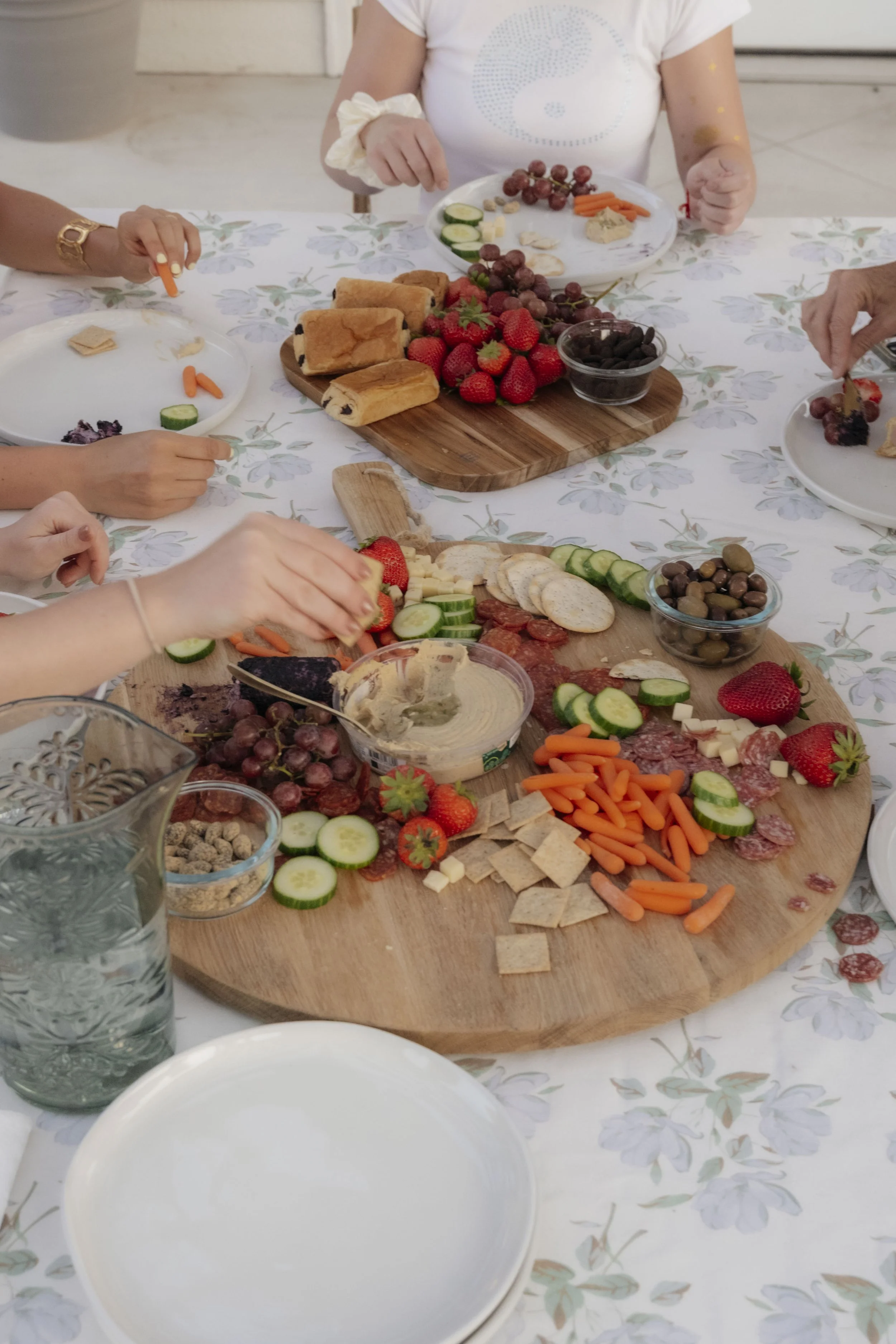 A buffet-style spread of various foods including strawberries, grapes, cucumber slices, carrots, crackers, cheese, and dips on a wooden platter, with people reaching for the food at a table covered with a floral tablecloth.