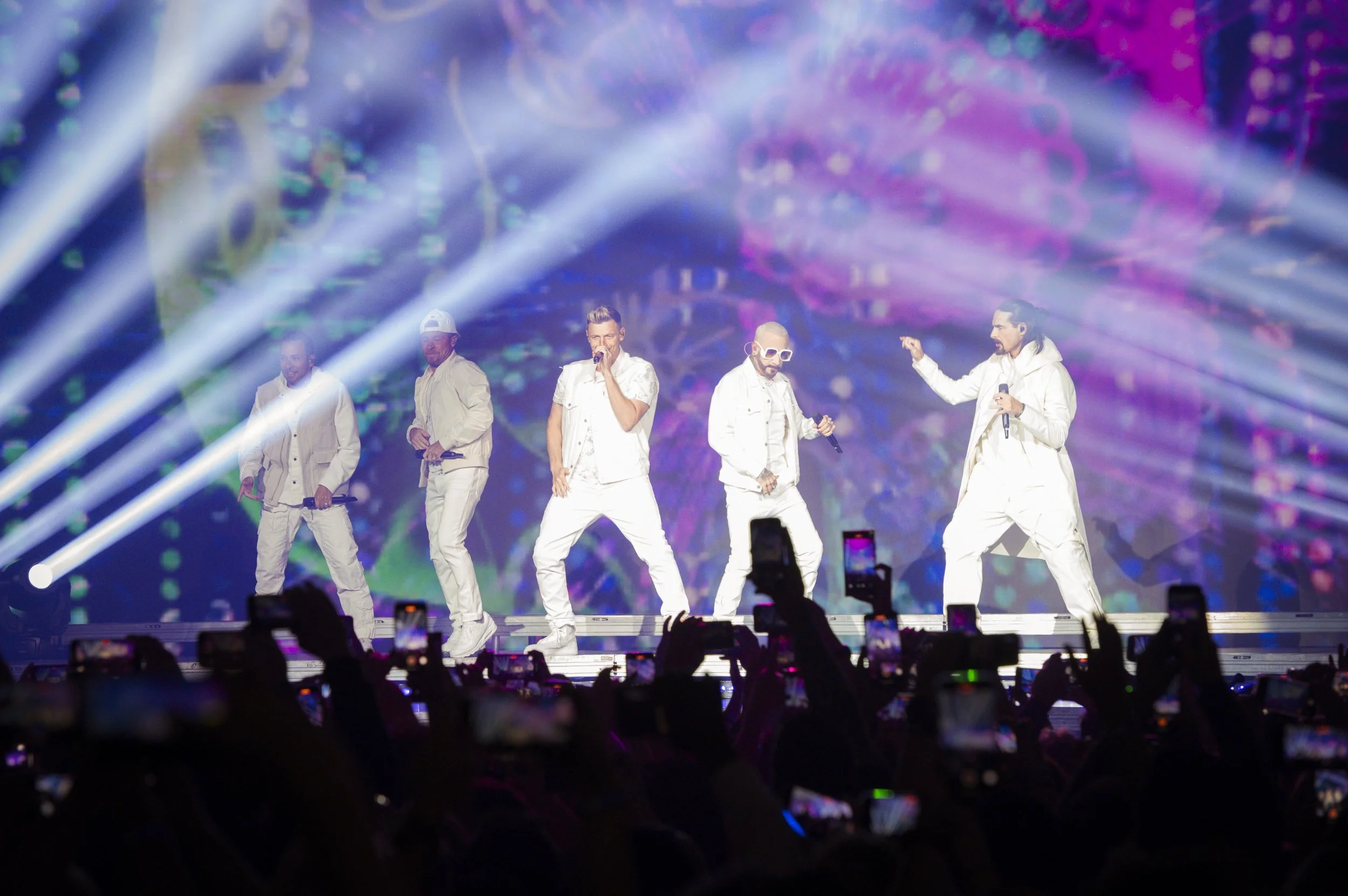 Backstreet Boys on stage with colorful lights and fog effects, wearing white outfits, with a crowd taking photos.