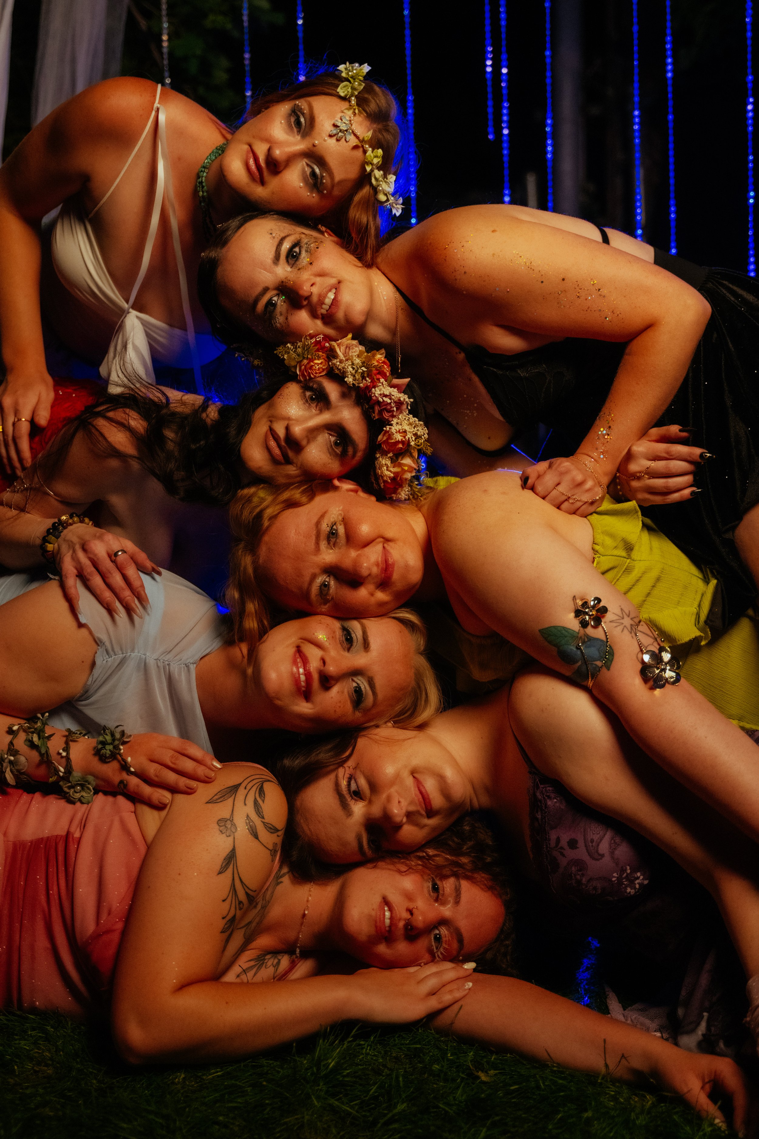 A group of women and one man lying closely together on the grass at night, with some wearing floral crowns, glitter makeup, and colorful clothing, in a festive outdoor setting with string lights in the background.