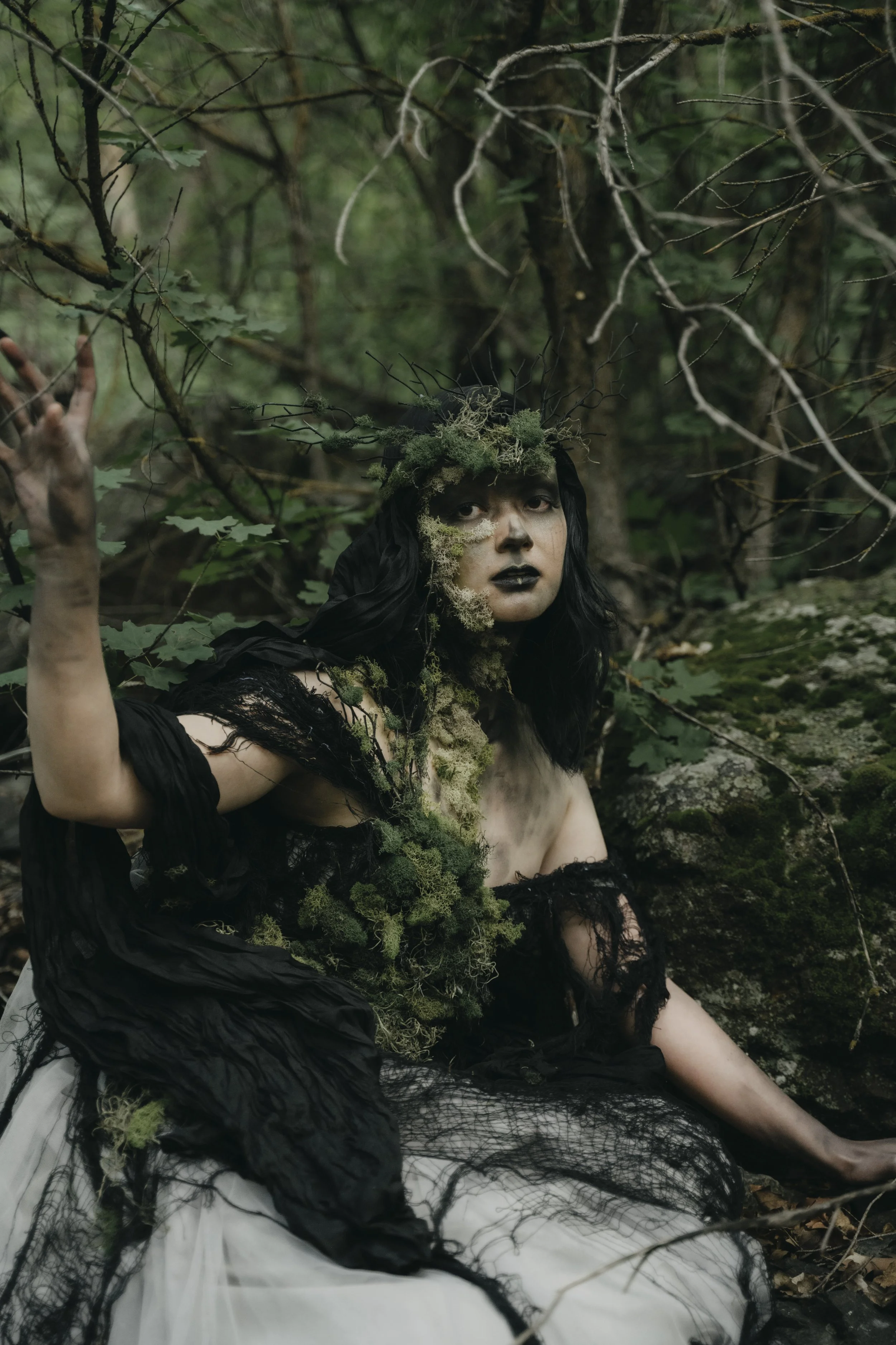Person in a dark, gothic costume with black hair, surrounded by forest, wearing plant-like decorations on their head and body, with a serious expression.