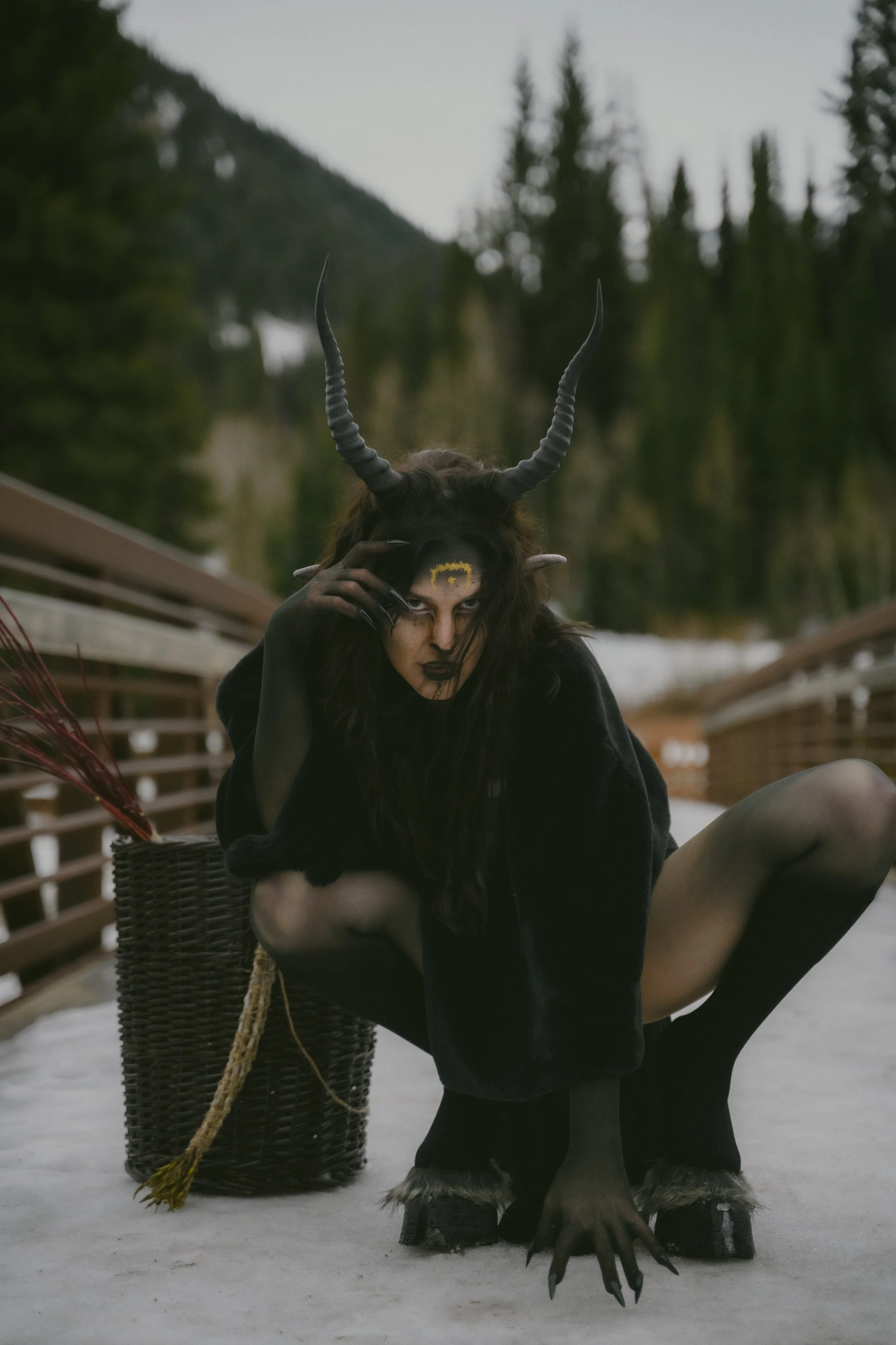 Person dressed as a creature with dark makeup, black costume, and horned headpiece, crouching on a snowy bridge with a forest background.