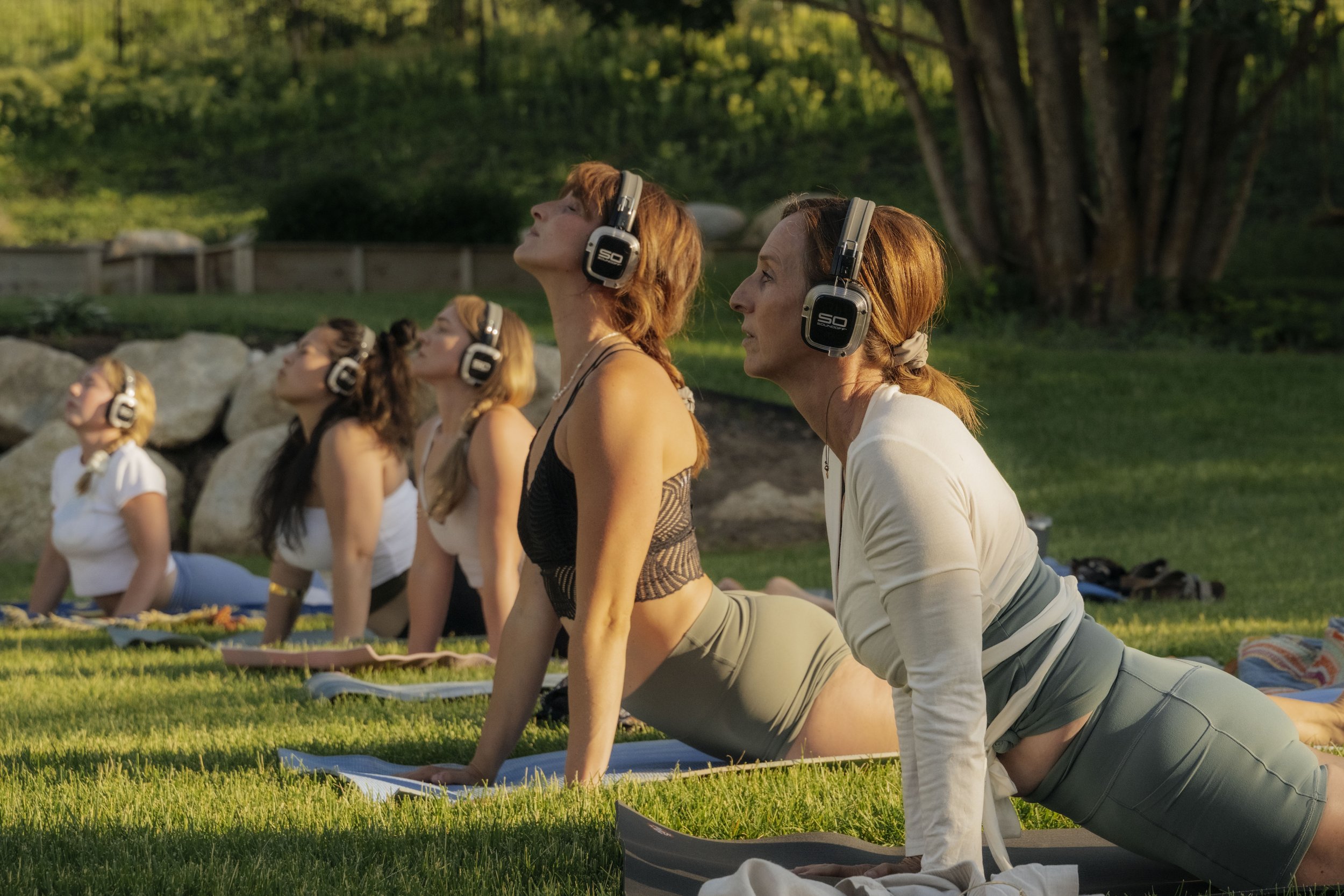 Group of women practicing yoga outdoors on grass during sunset, wearing headphones, in a yoga pose with upward gaze.