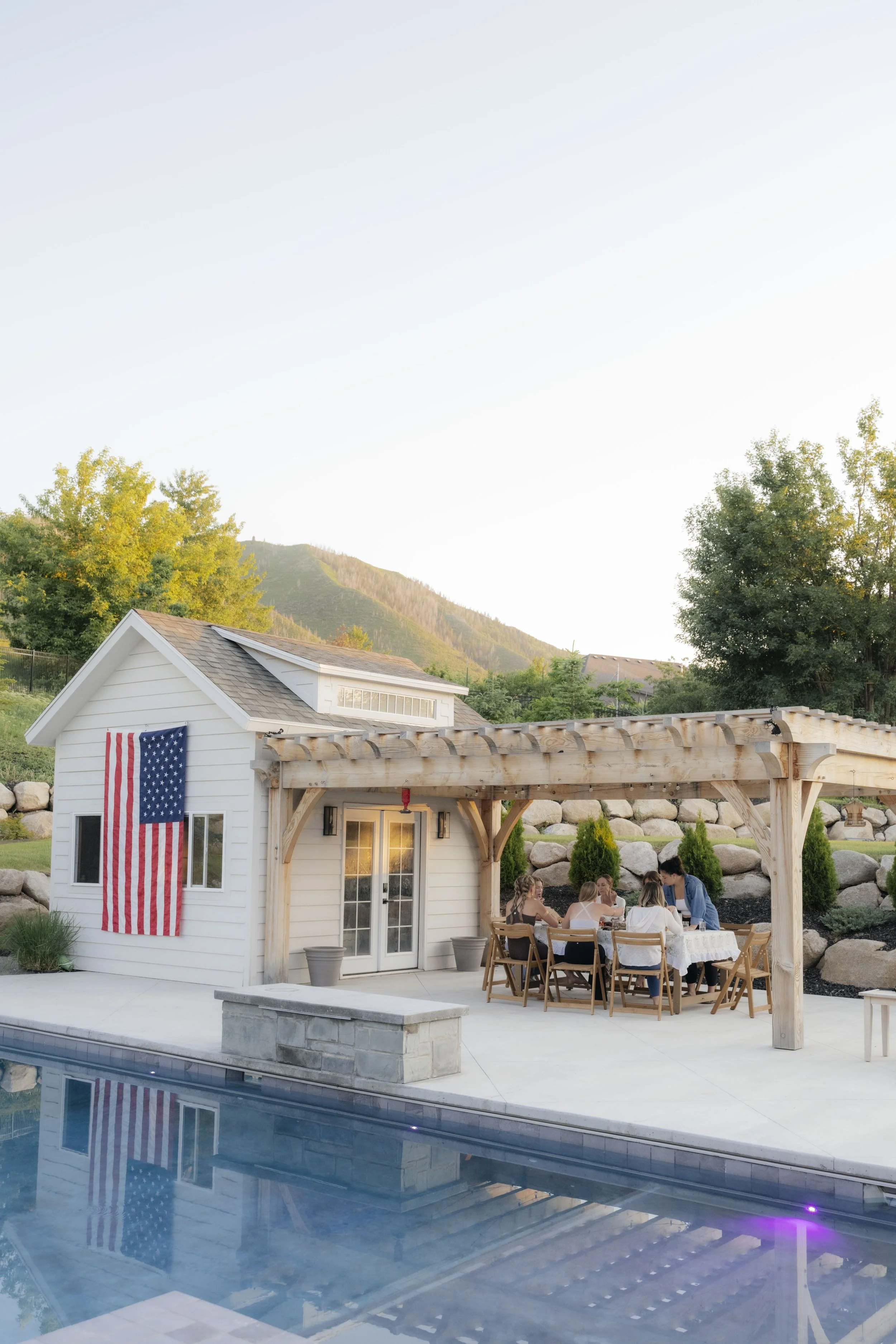 A group of people gathered around a table on a patio next to a pool, with a small white house decorated with an American flag in the background, surrounded by trees and mountains.